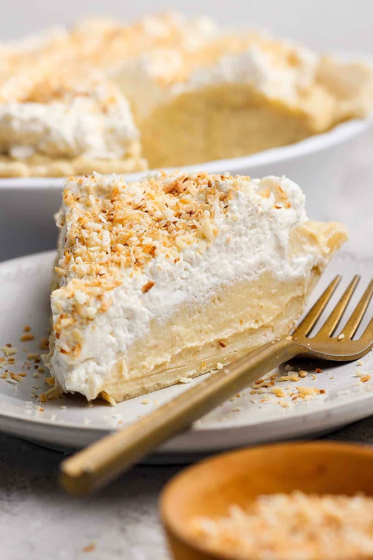 A slice of coconut cream pie topped with whipped cream and toasted coconut flakes on a plate with a gold fork.