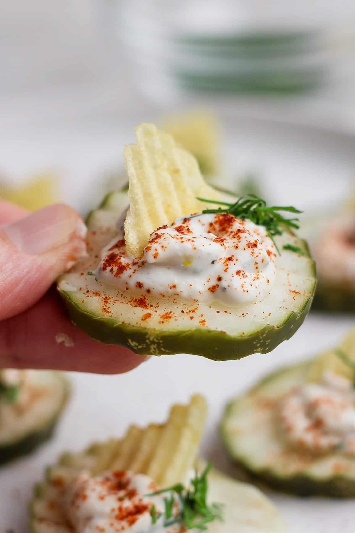 A hand holding a sliced cucumber topped with a dollop of cream, a sprinkle of paprika, a piece of ridged potato chip, and a sprig of dill.