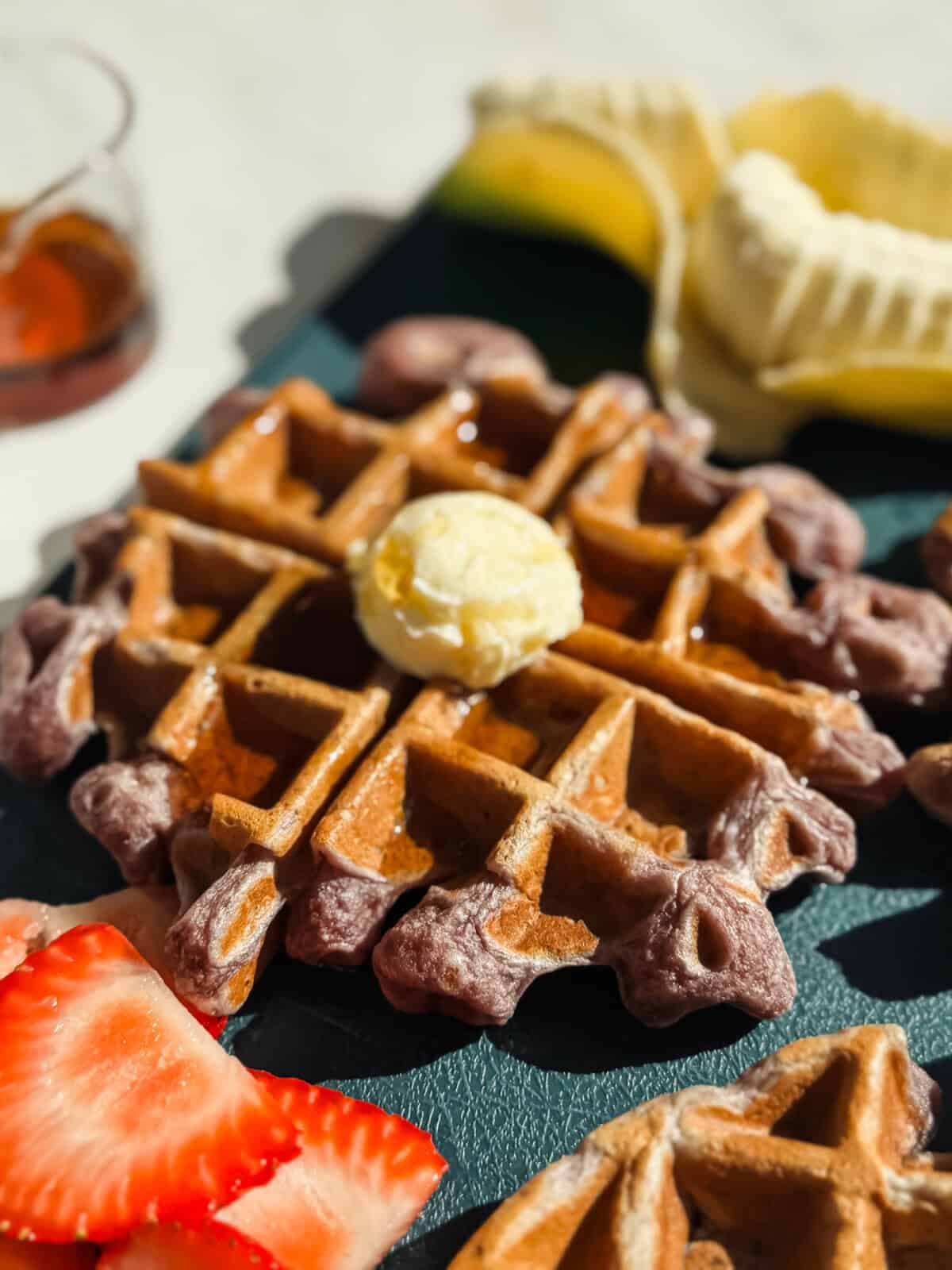 A close-up of a waffle topped with a scoop of butter and syrup, with sliced strawberries and a partially peeled banana in the background.