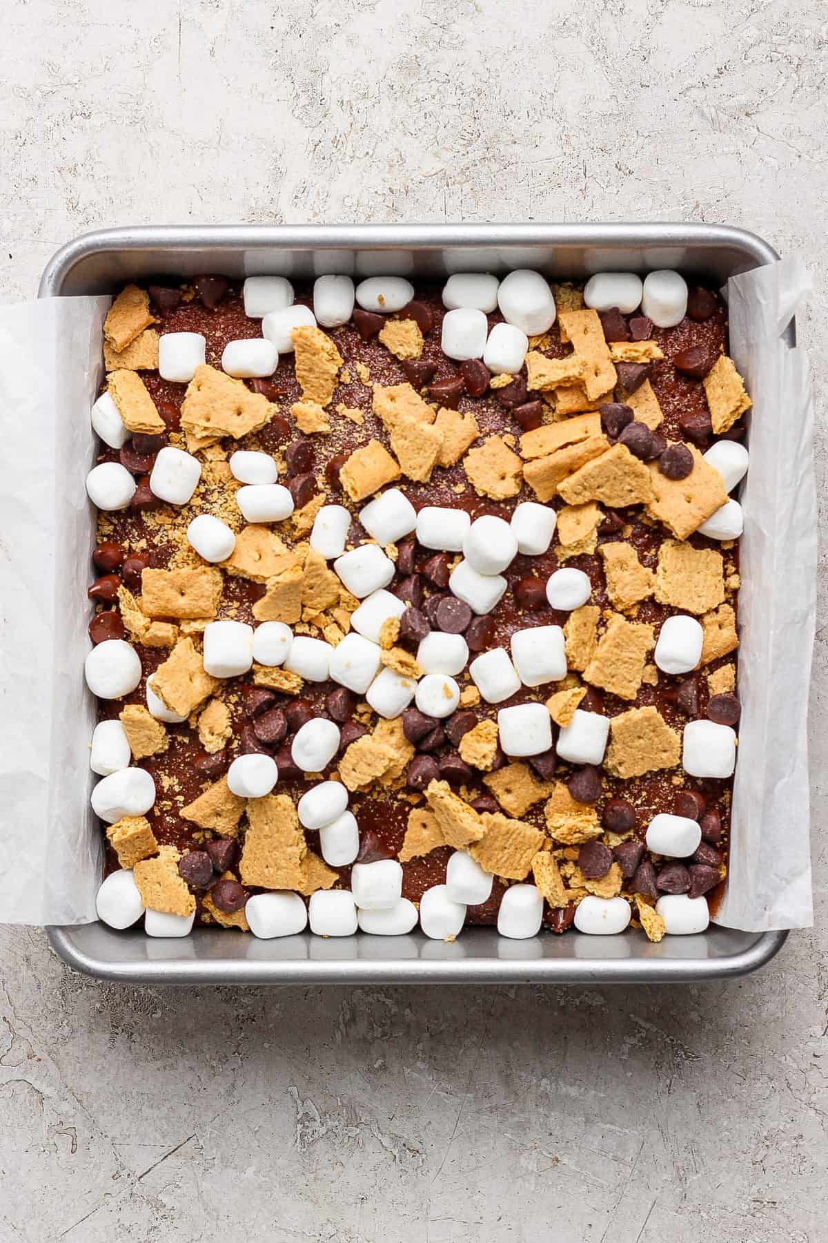 A square baking pan lined with parchment paper, filled with a mixture of mini marshmallows, chocolate chips, and broken graham crackers sits atop a rich s'mores brownies base.