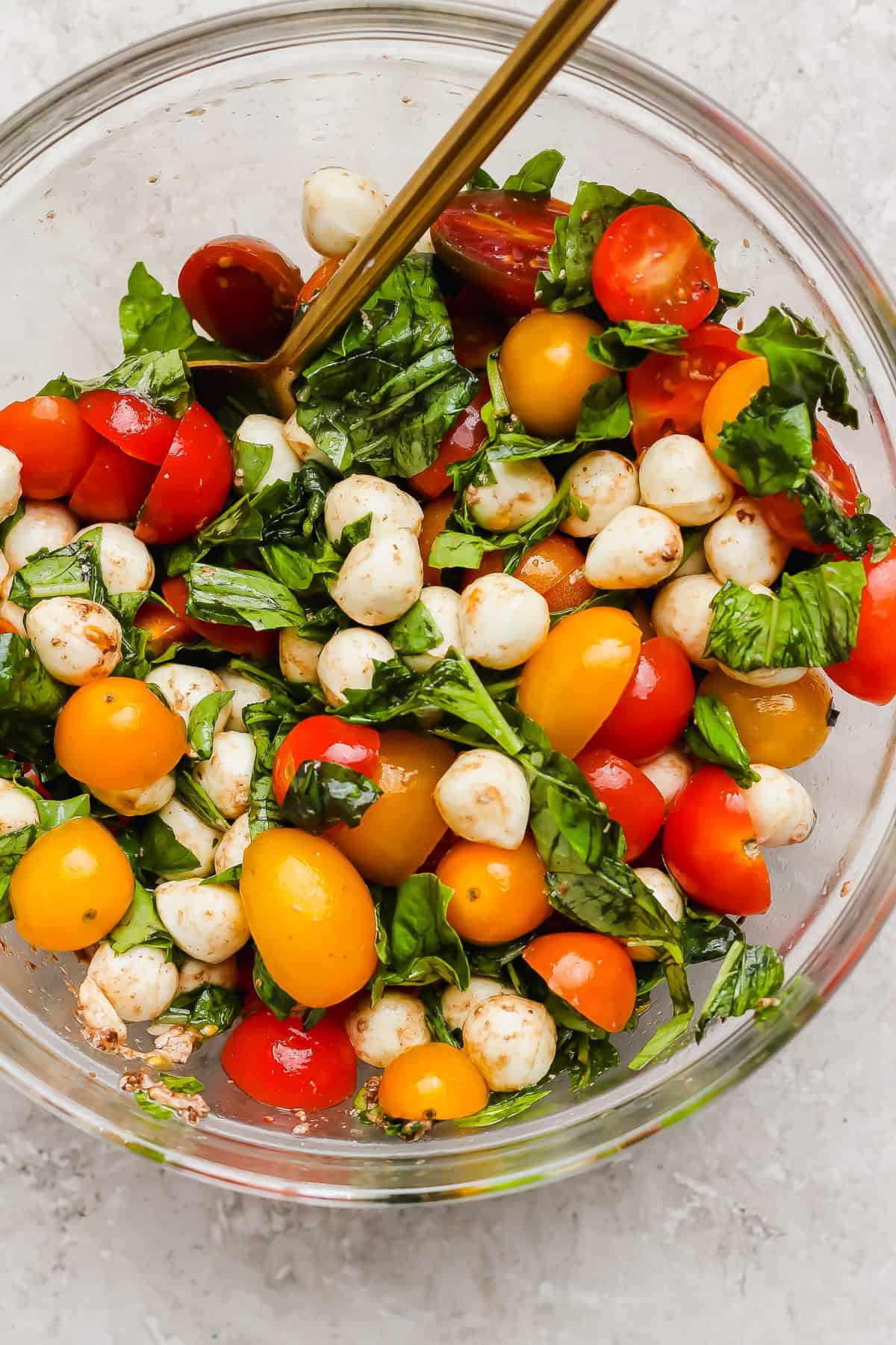 A clear glass bowl filled with cherry tomatoes, mozzarella balls, fresh basil leaves, and a spoon, on a light surface.