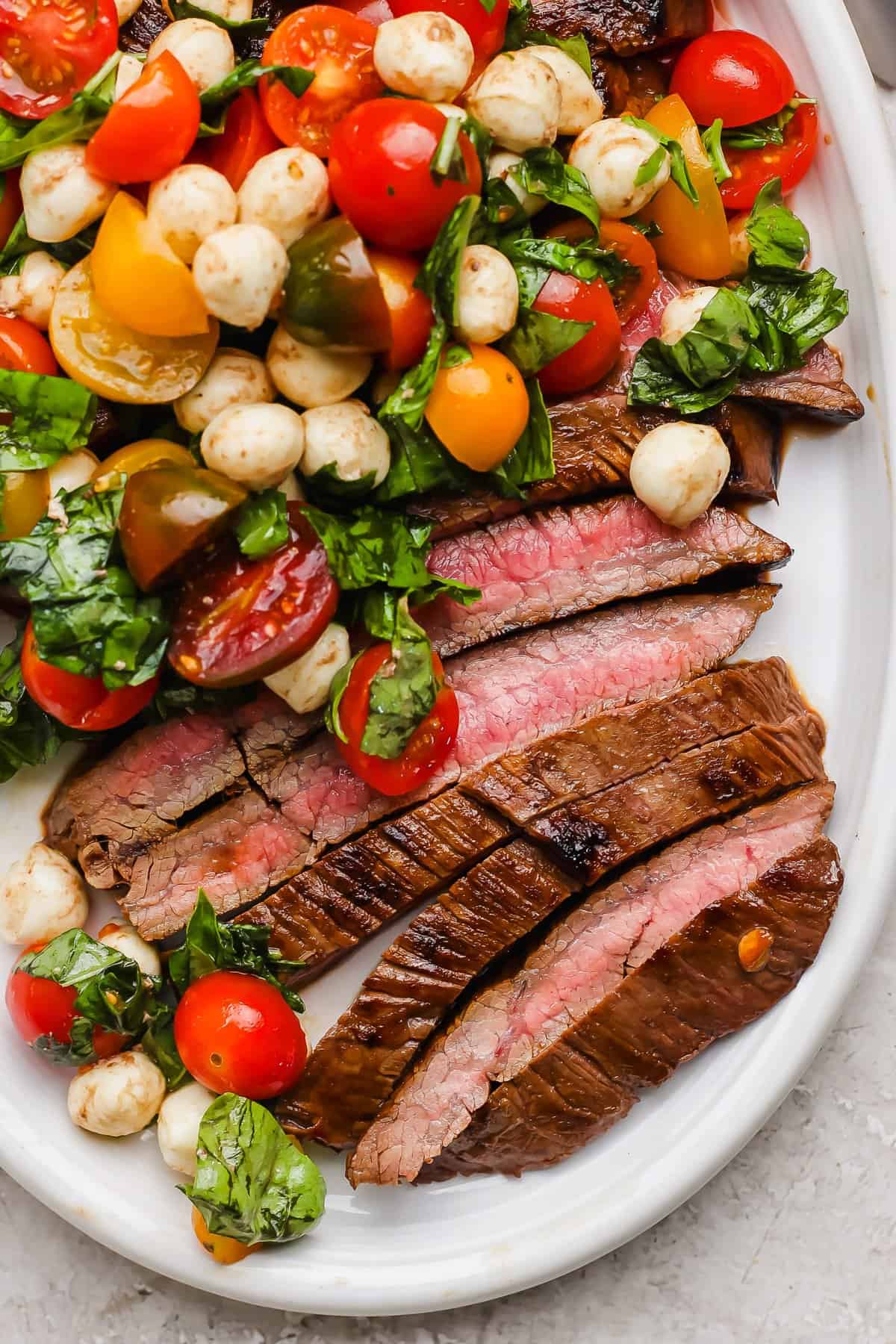 Sliced medium-rare steak served with a salad of cherry tomatoes, mozzarella balls, and fresh basil on a white plate.