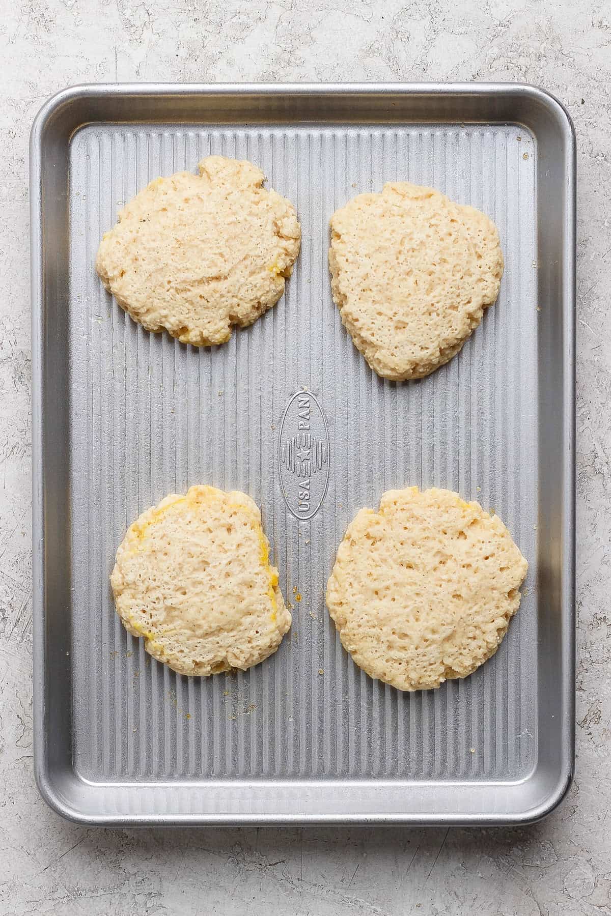 Four unbaked biscuit dough rounds are spaced evenly on a metal baking sheet placed on a light-colored surface.