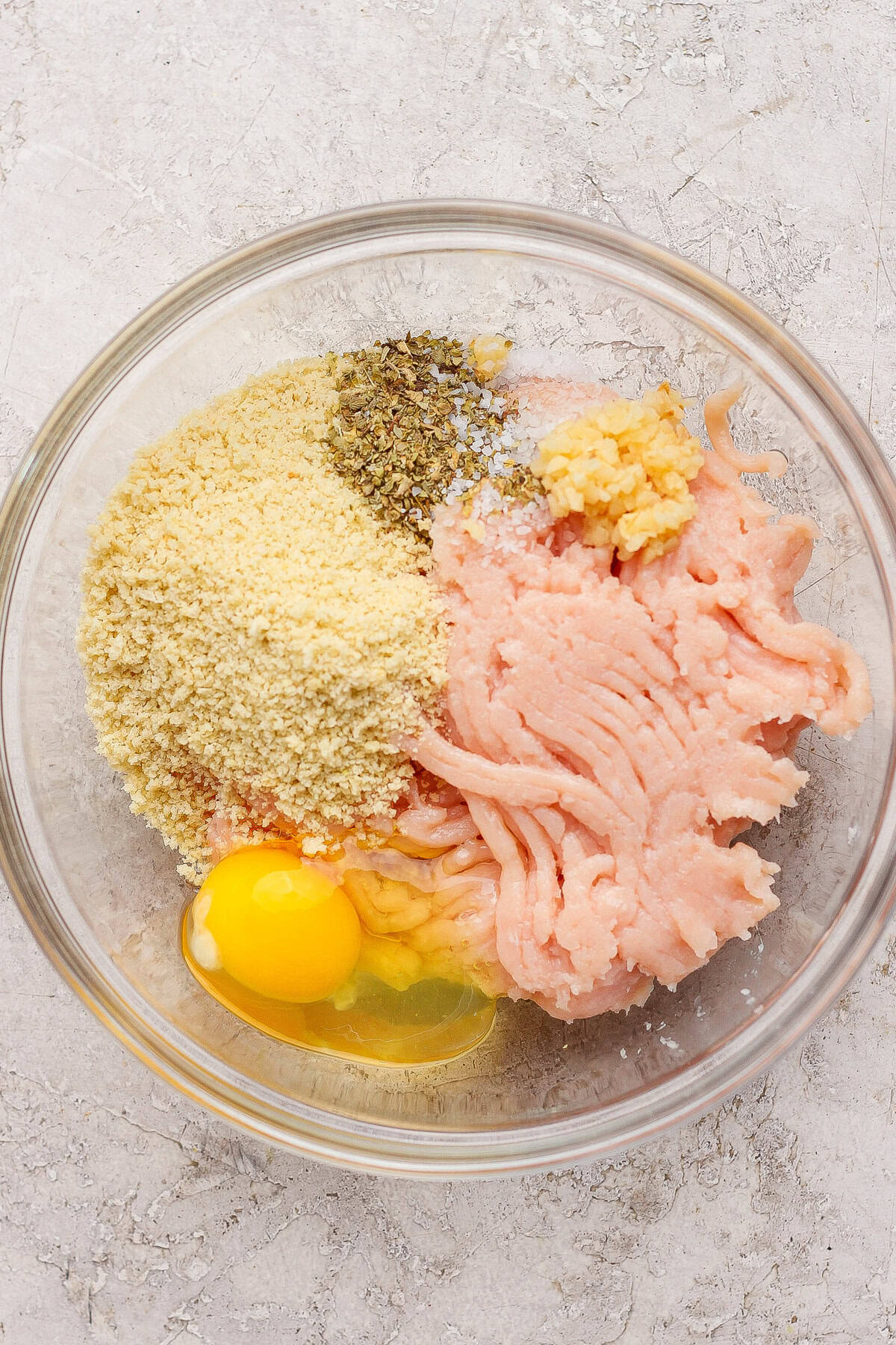 A glass bowl containing ground chicken, a cracked egg, breadcrumbs, minced garlic, salt, and dried herbs on a light textured surface.