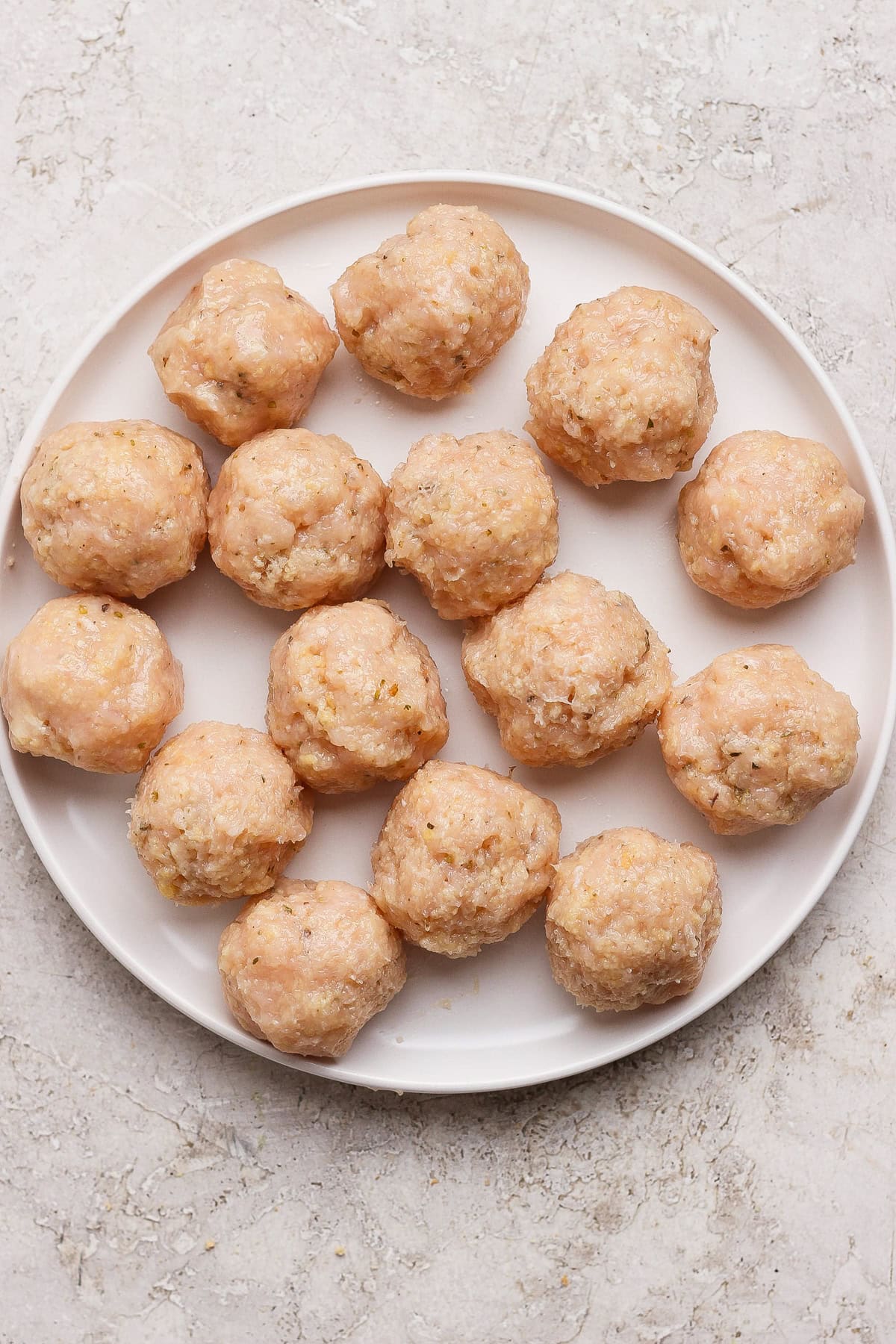 A white plate with sixteen raw, uncooked meatballs arranged in a circular pattern on a light textured surface.
