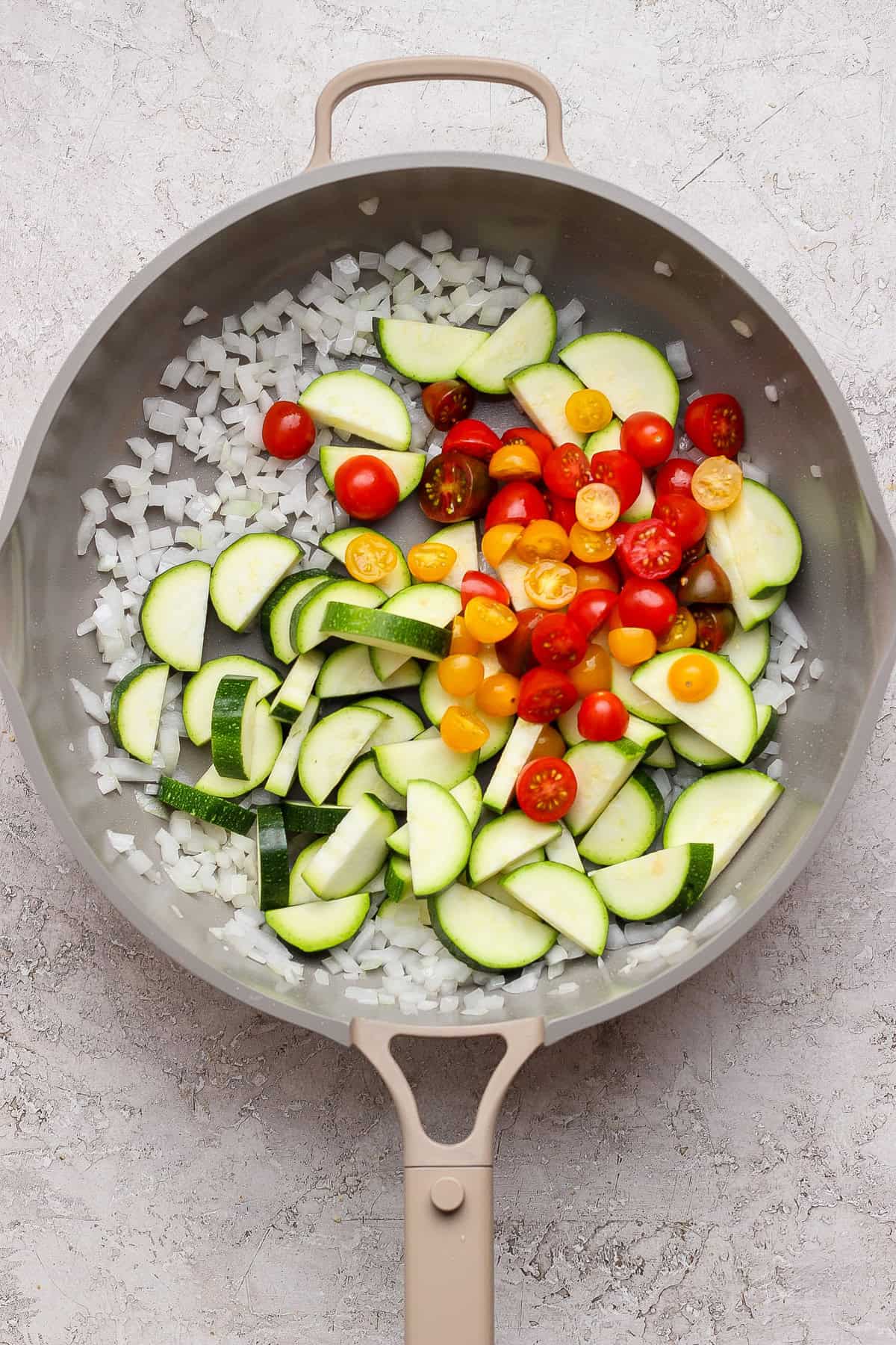 A frying pan contains chopped onions, sliced zucchini, and halved cherry tomatoes on a light textured surface.