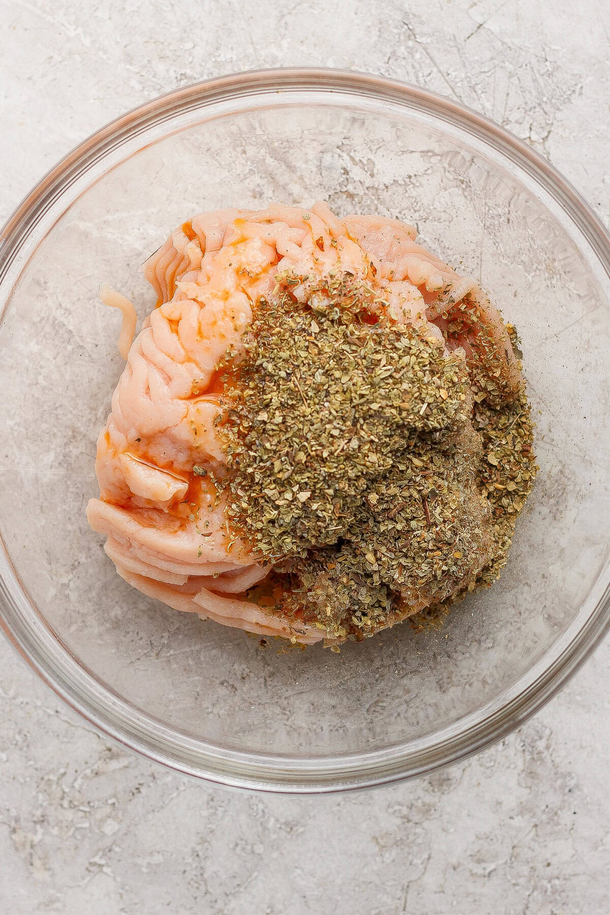 Raw ground meat topped with dried herbs and spices in a clear glass bowl on a light textured surface.