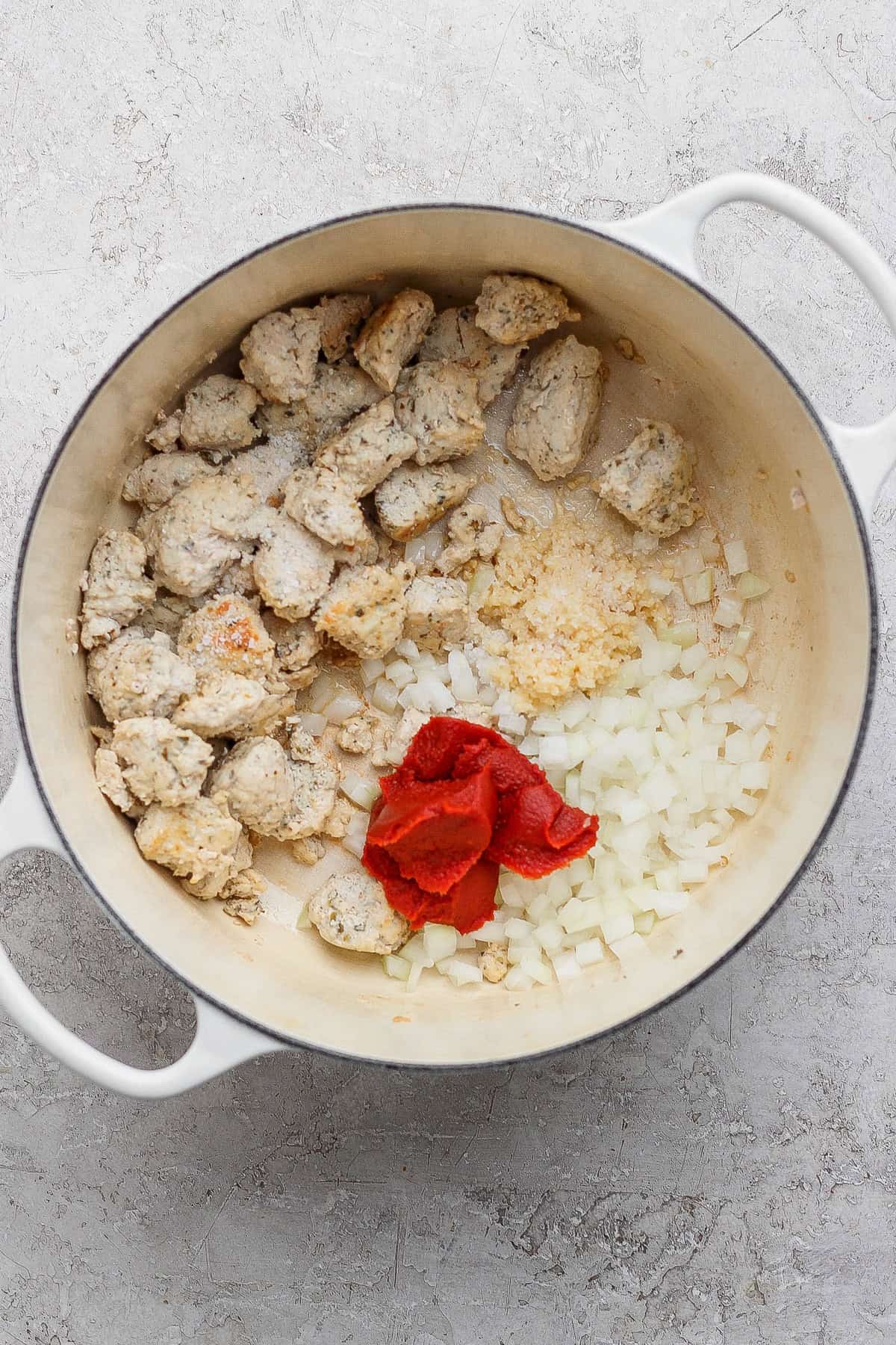 A white pot contains cooked chunks of sausage, chopped onions, minced garlic, and tomato paste on a light countertop.