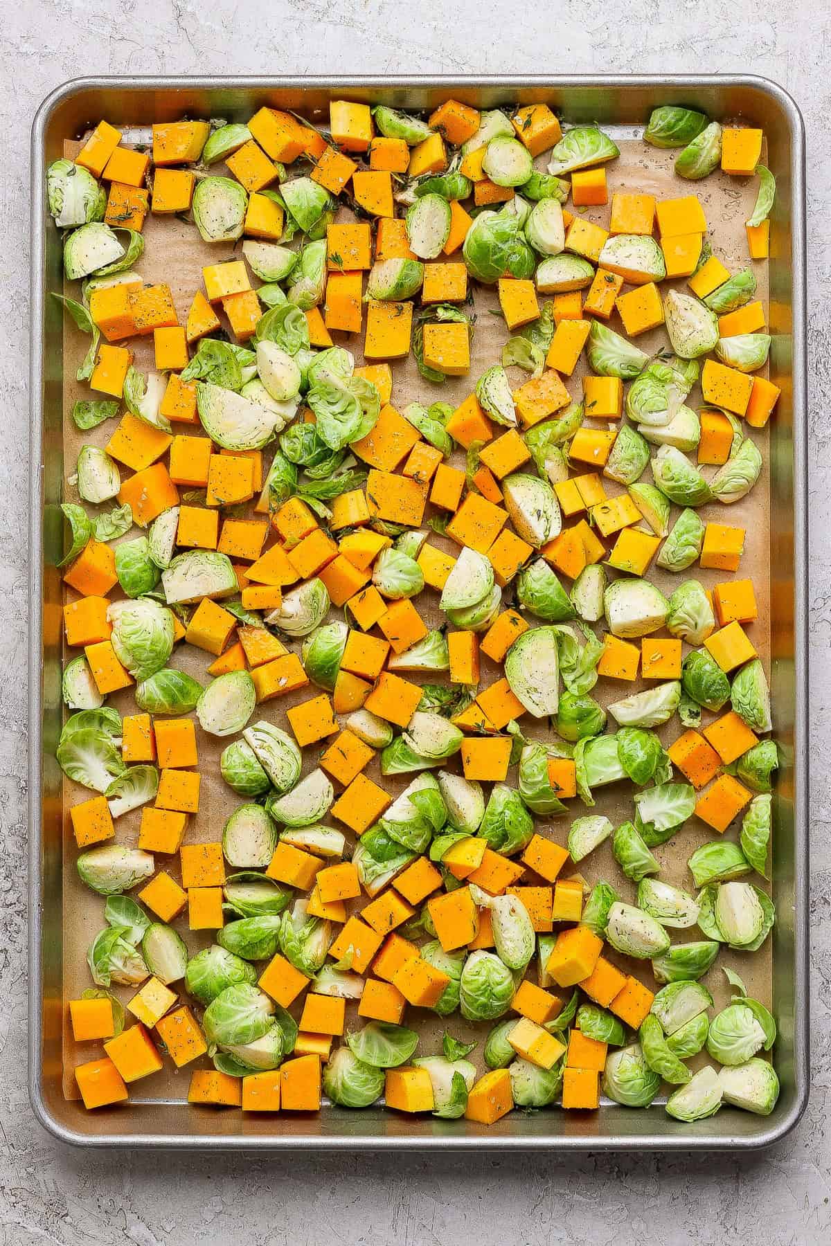 Cubed butternut squash and halved Brussels sprouts spread on a parchment-lined baking sheet, seasoned with salt, pepper, and herbs—perfect for adding to your favorite couscous salad recipe after roasting.