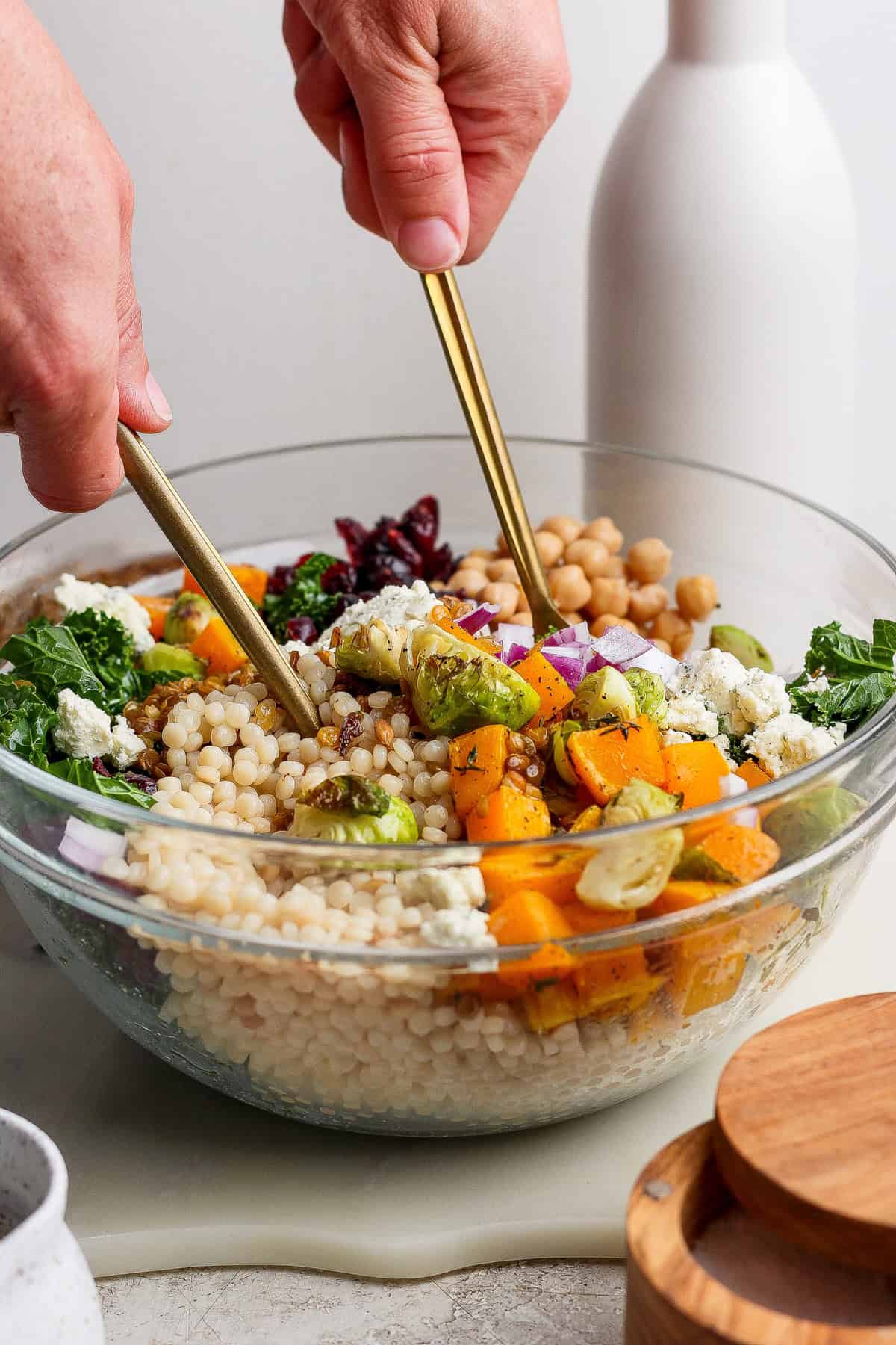 A person tossing a couscous salad recipe in a glass bowl with chickpeas, roasted vegetables, kale, red onion, cranberries, and cheese using two gold utensils.