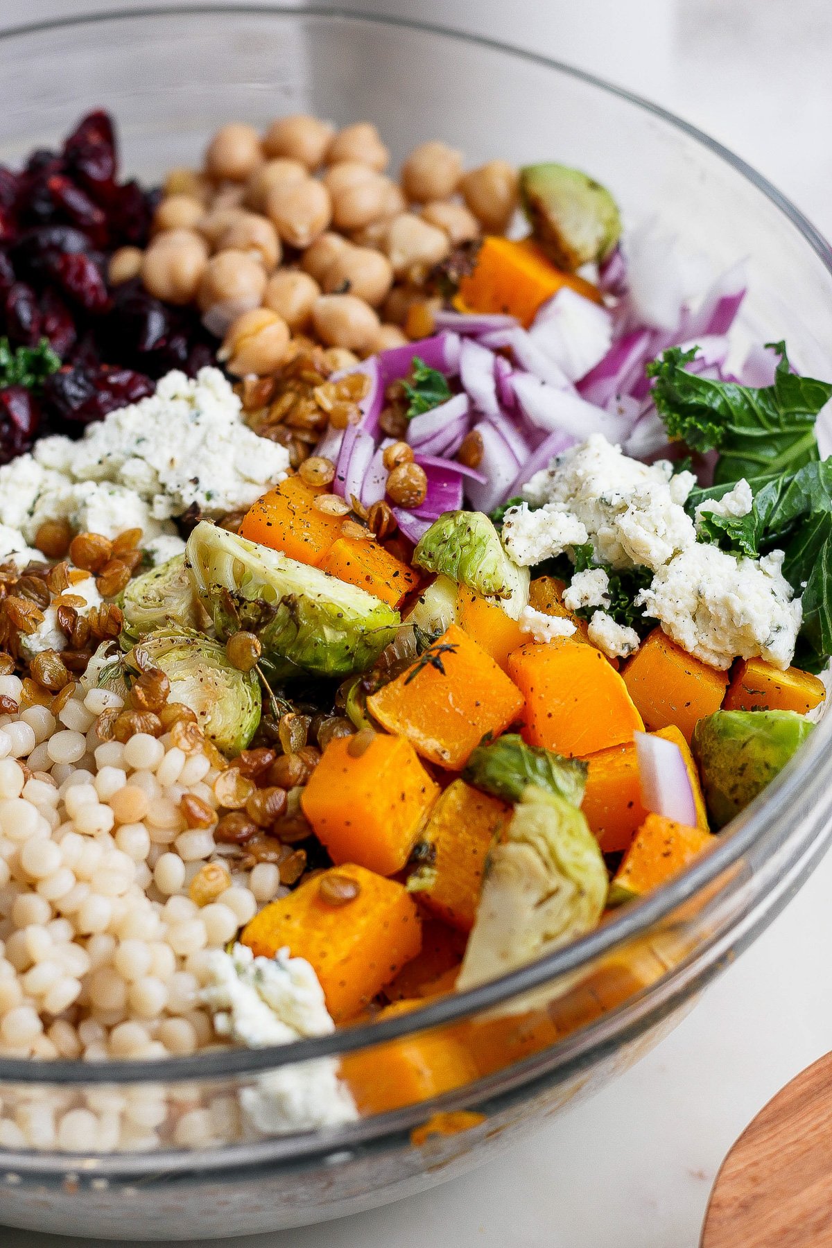 A glass bowl filled with a vibrant couscous salad recipe featuring chickpeas, dried cranberries, red onions, kale, roasted squash, Brussels sprouts, lentils, and crumbled cheese.