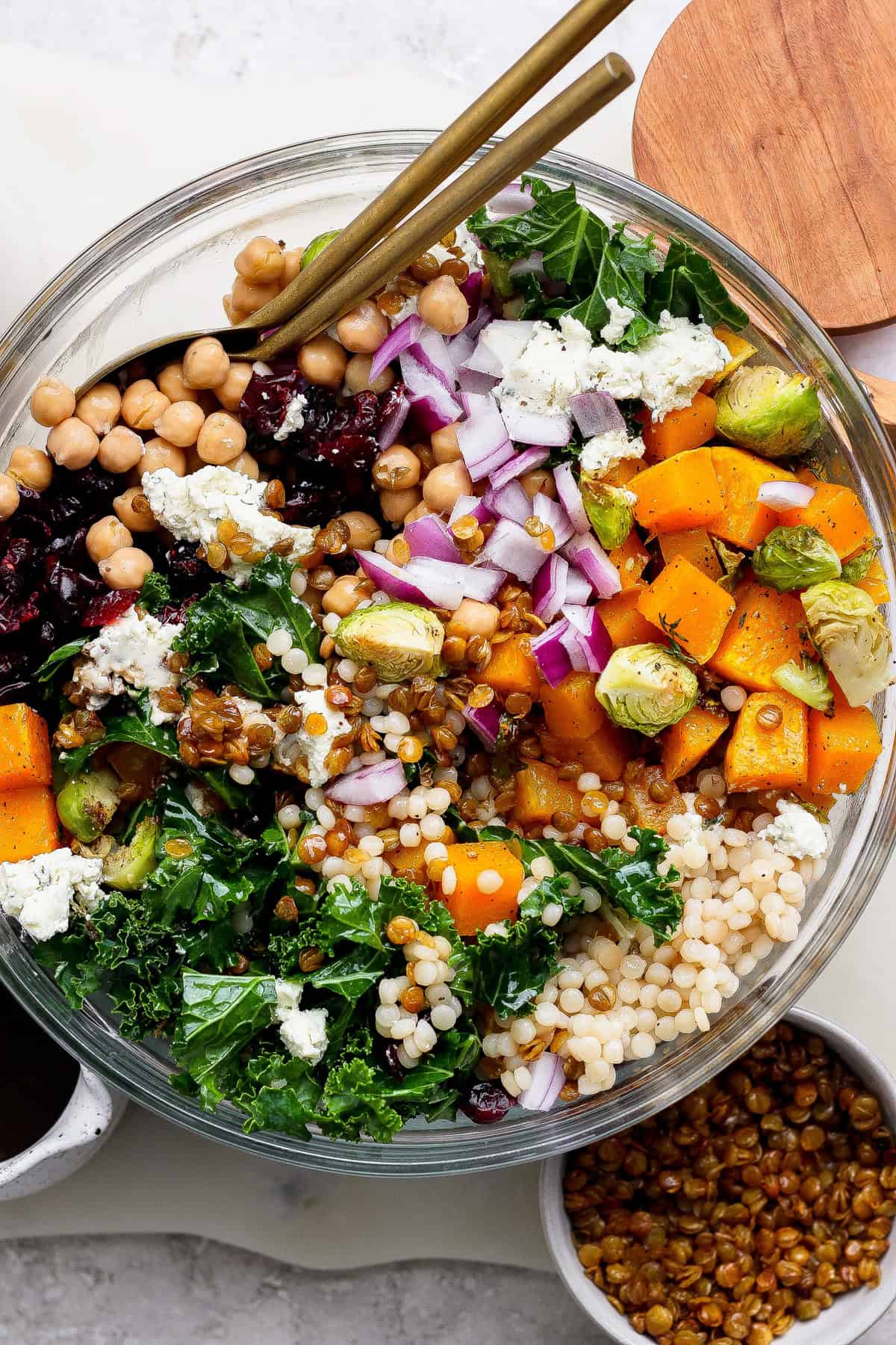 A vibrant couscous salad recipe featuring kale, chickpeas, roasted butternut squash, Brussels sprouts, red onion, blue cheese, dried cranberries, and lentils in a glass bowl with gold utensils on top.