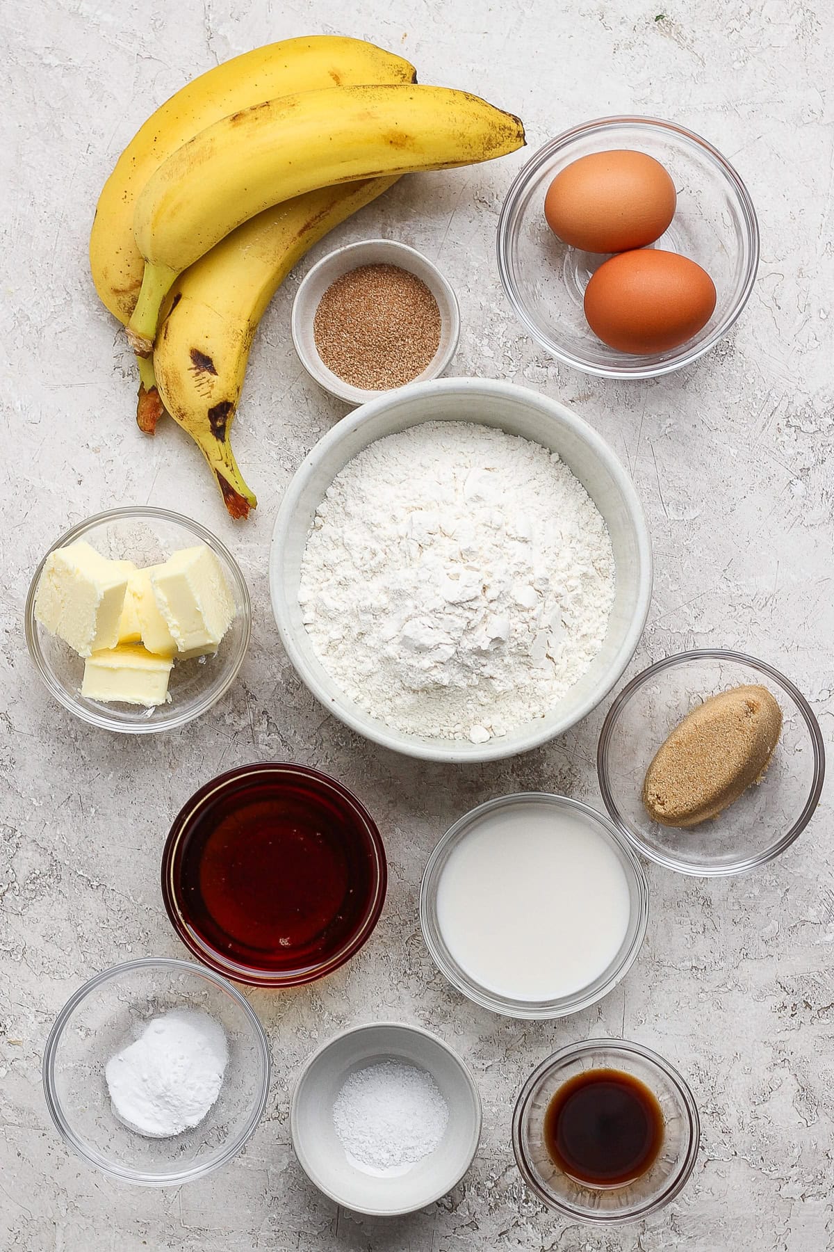Banana bread recipe essentials: bananas, eggs, flour, sugar, butter, milk, brown sugar, baking powder, vanilla extract, salt, and cinnamon are beautifully arranged in bowls on a light surface.