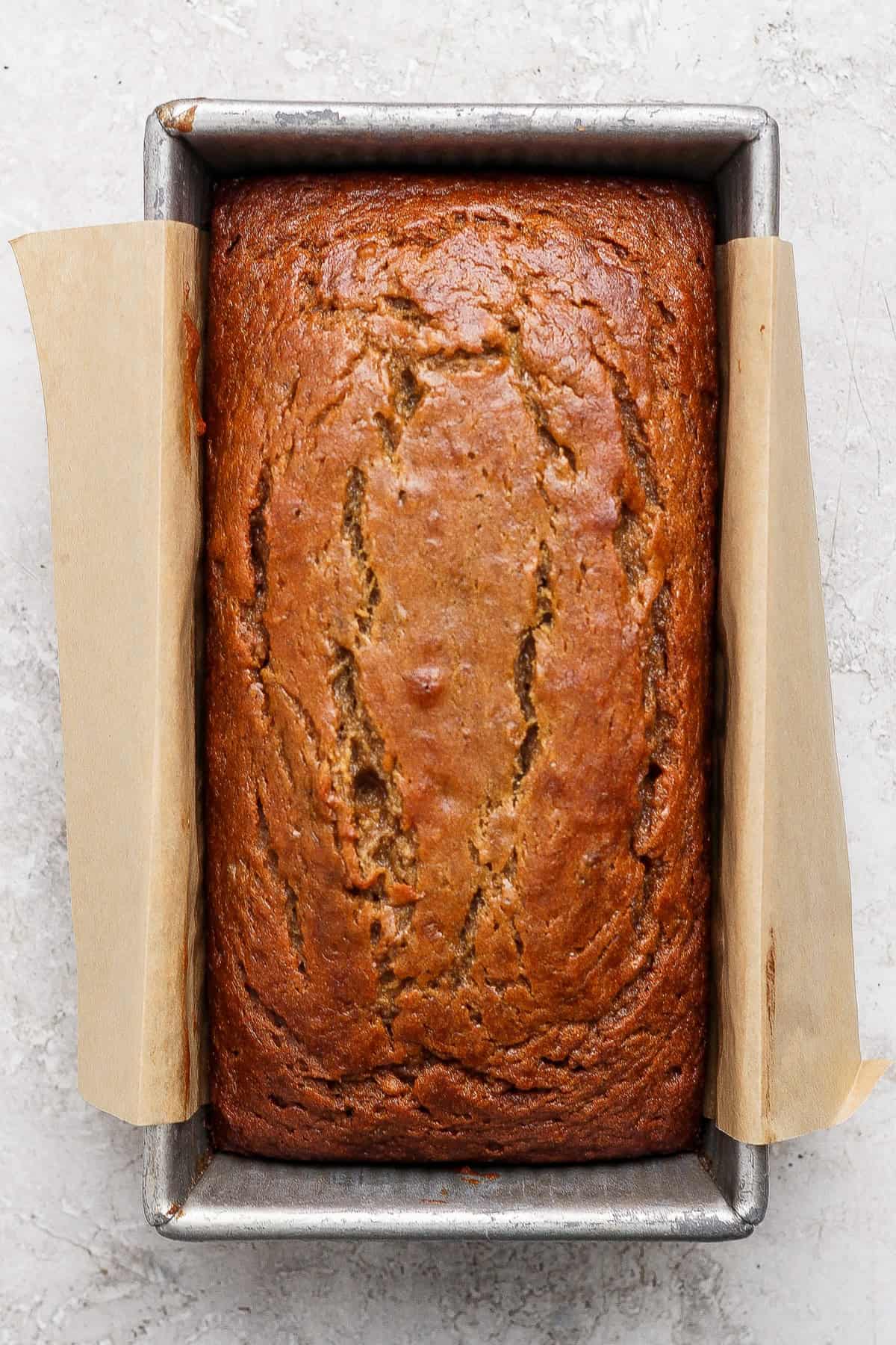 A loaf of banana bread from a classic banana bread recipe sits in a rectangular metal pan lined with parchment paper, shown from above on a light textured surface.