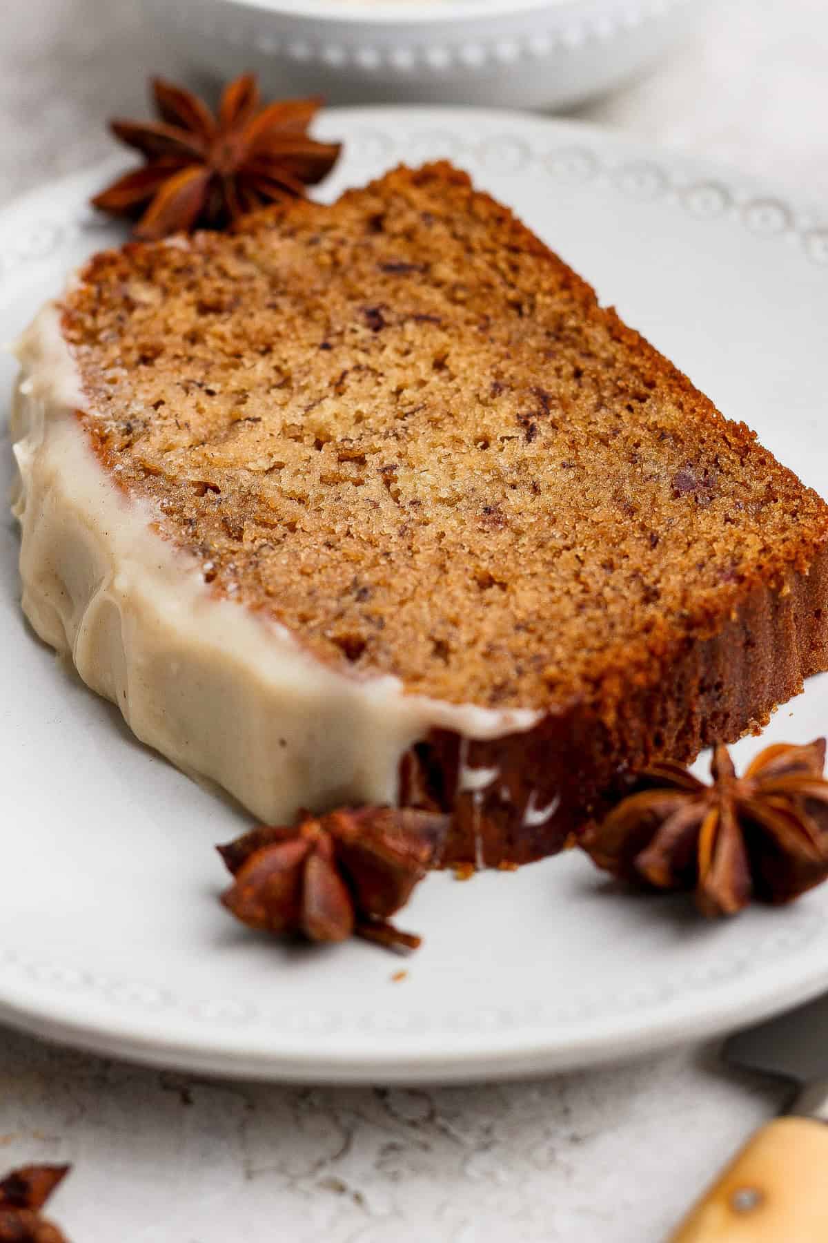 A slice of spiced banana bread loaf with white glaze on one edge, garnished with whole star anise, sits elegantly on a white plate—a delicious twist on the classic banana bread recipe.