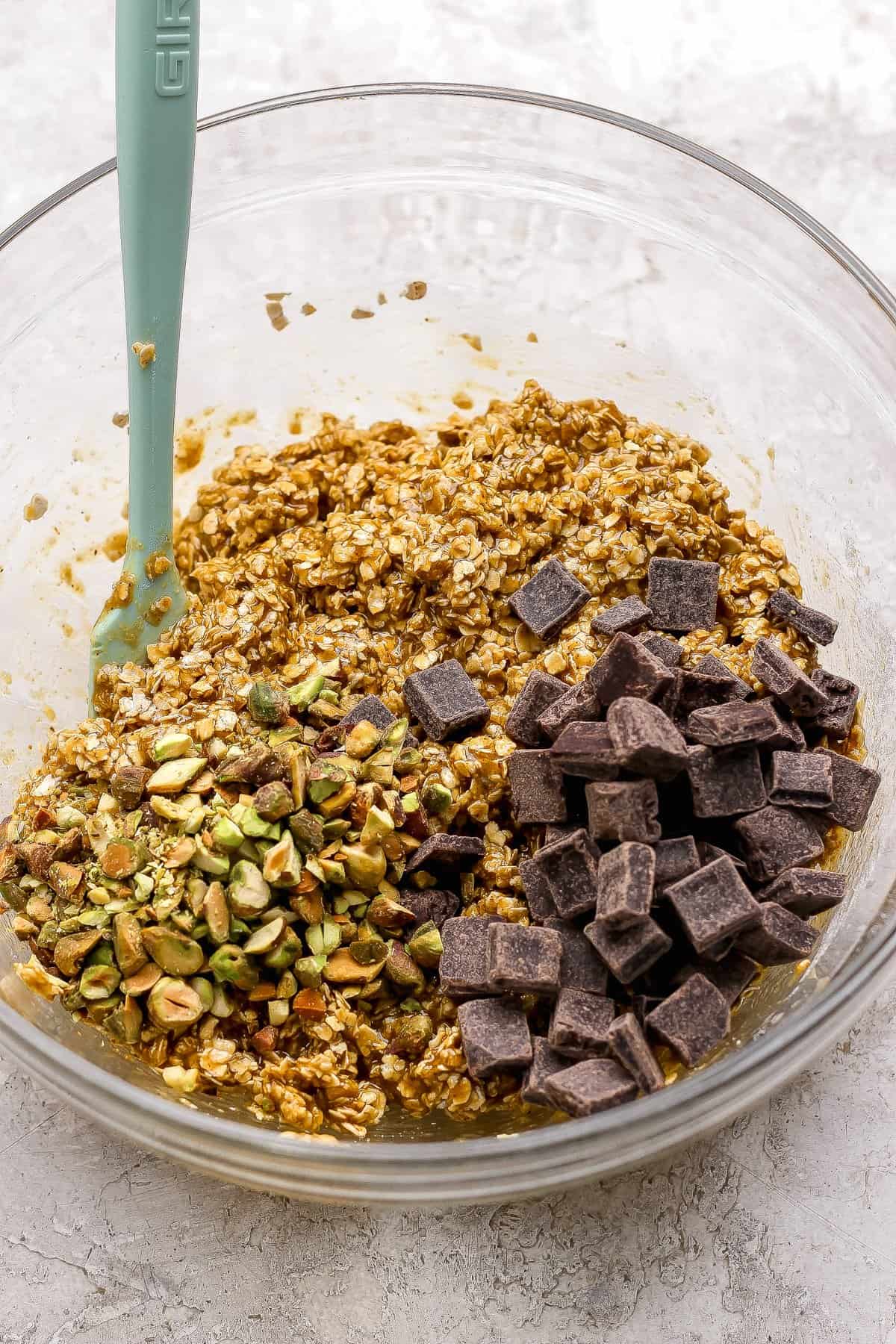 A glass bowl containing oat mixture with chopped pistachios and dark chocolate chunks, and a green spatula resting inside.