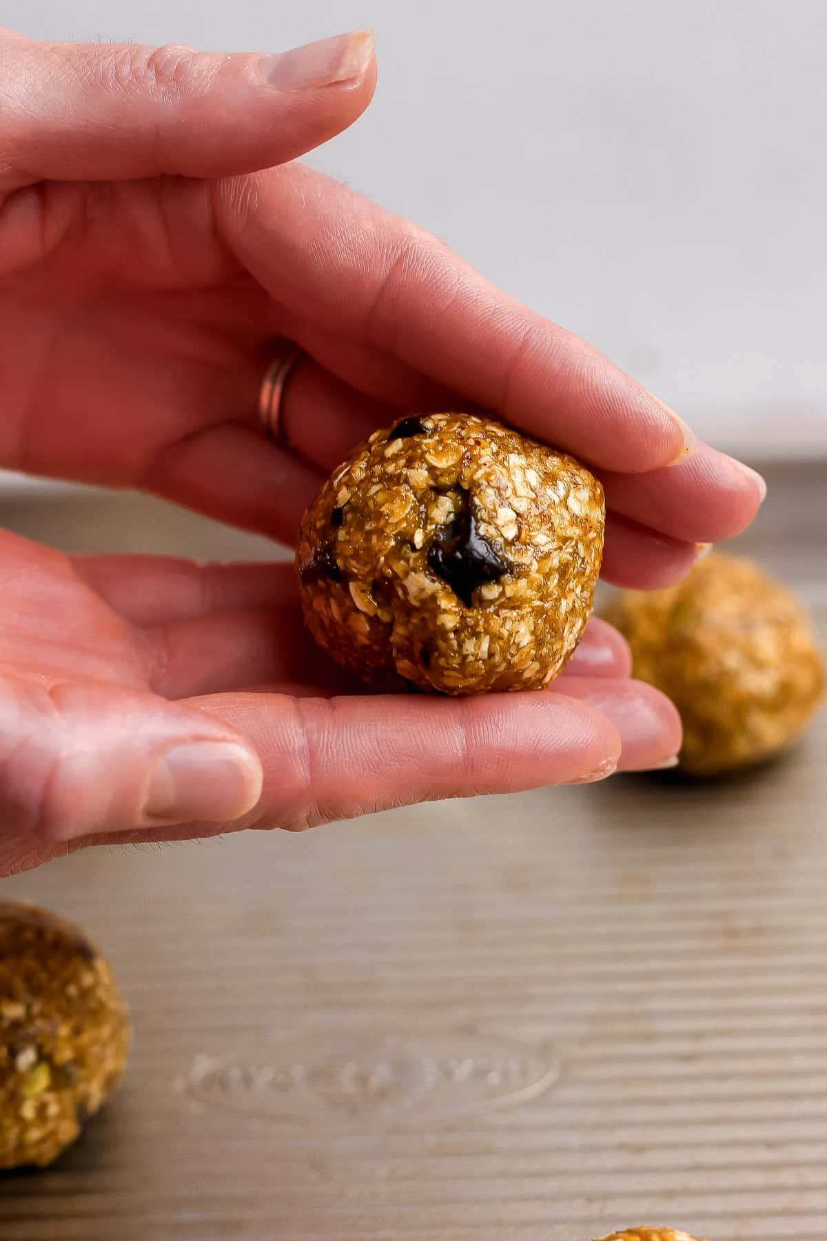 A hand holding a round, oat-based energy bite with a visible chocolate chip, above a metal baking tray with more energy bites in the background.