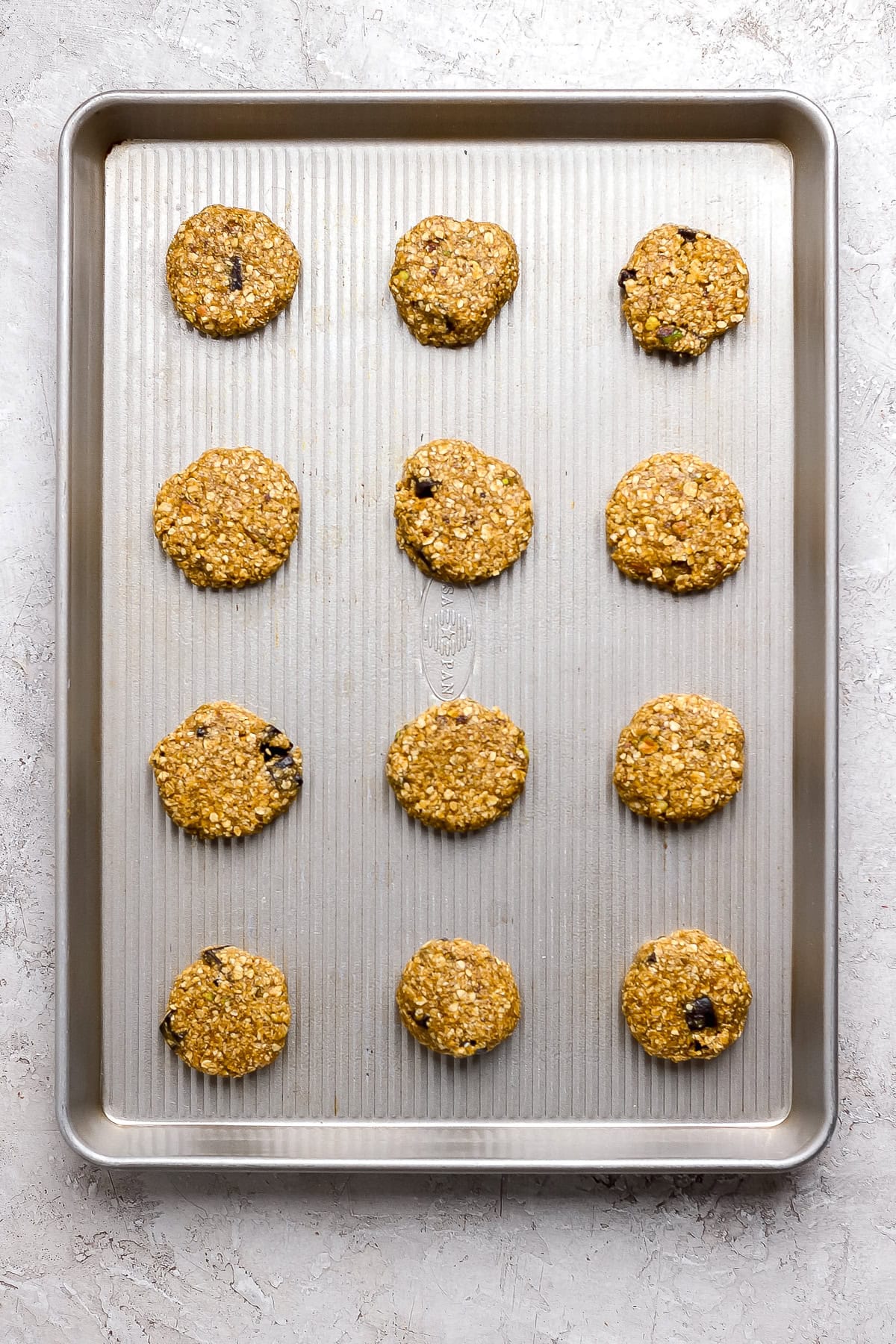 Twelve unbaked oatmeal cookies with chocolate chips are evenly spaced on a metal baking sheet, ready to be baked.