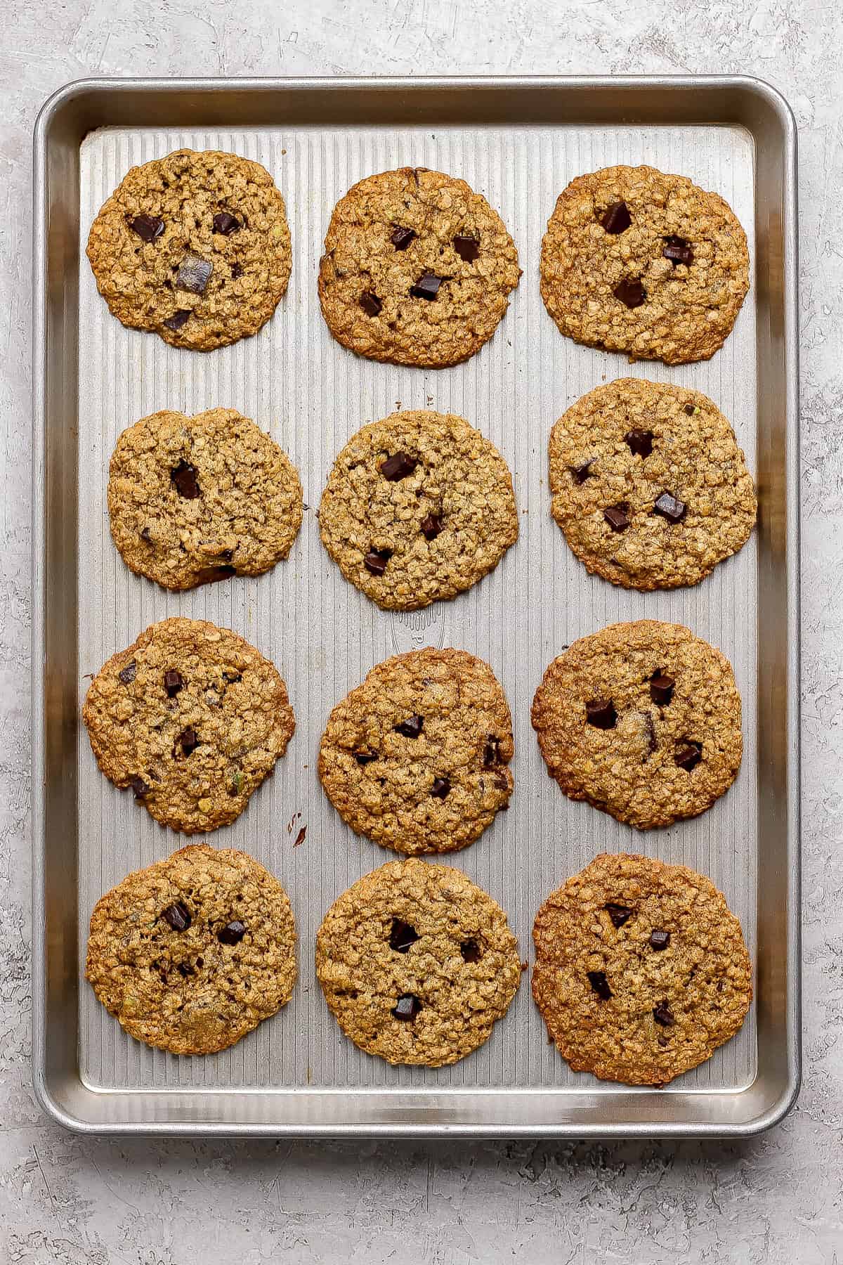 A baking sheet holds twelve baked oatmeal chocolate chip cookies arranged in three rows of four on a light-colored surface.