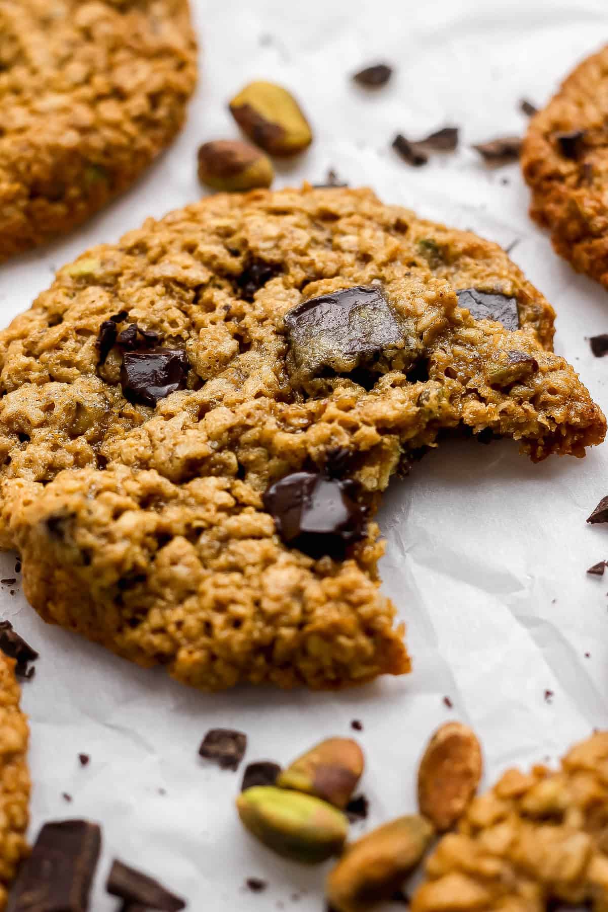 A close-up of an oatmeal chocolate chip cookie with a bite taken out, surrounded by chocolate chunks and pistachios on parchment paper.