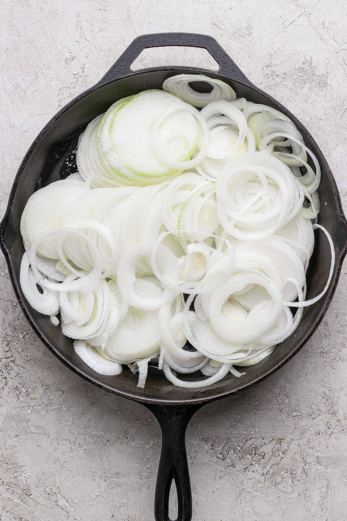 A cast iron skillet filled with raw, sliced white onions, arranged in overlapping rings on a light textured surface.