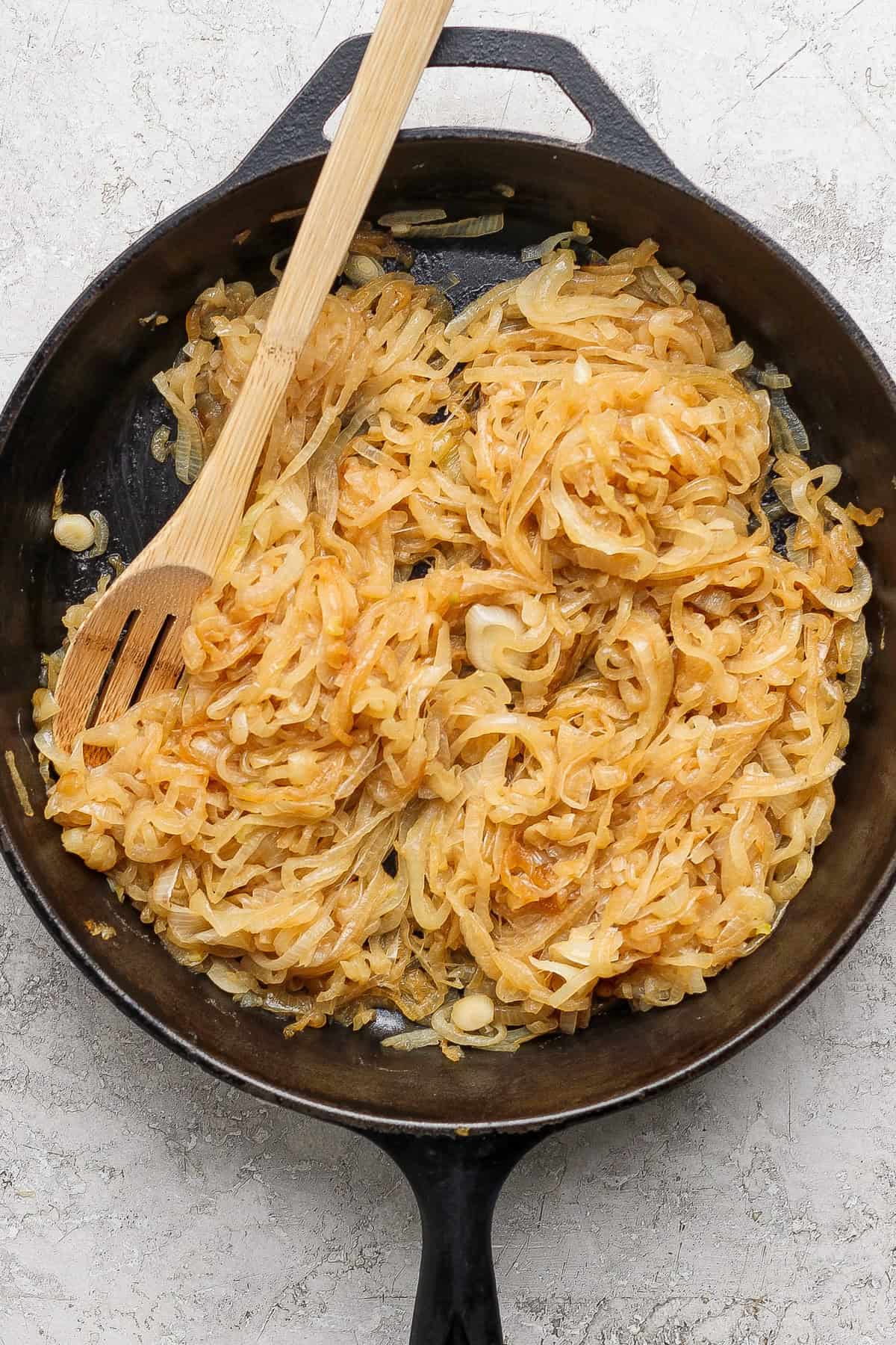 Sliced onions caramelizing in a black cast iron skillet with a wooden spoon resting on top.