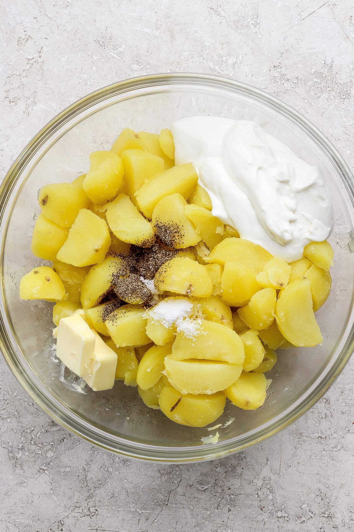 A glass bowl containing boiled, sliced potatoes, a pat of butter, a dollop of sour cream, salt, and black pepper on a light surface.