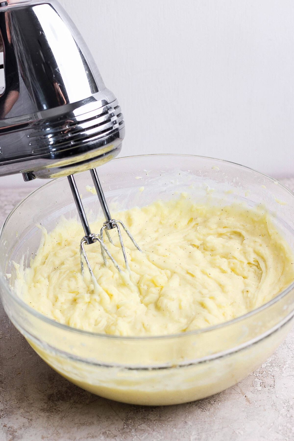 A handheld electric mixer blending creamy batter in a clear glass bowl on a light counter.