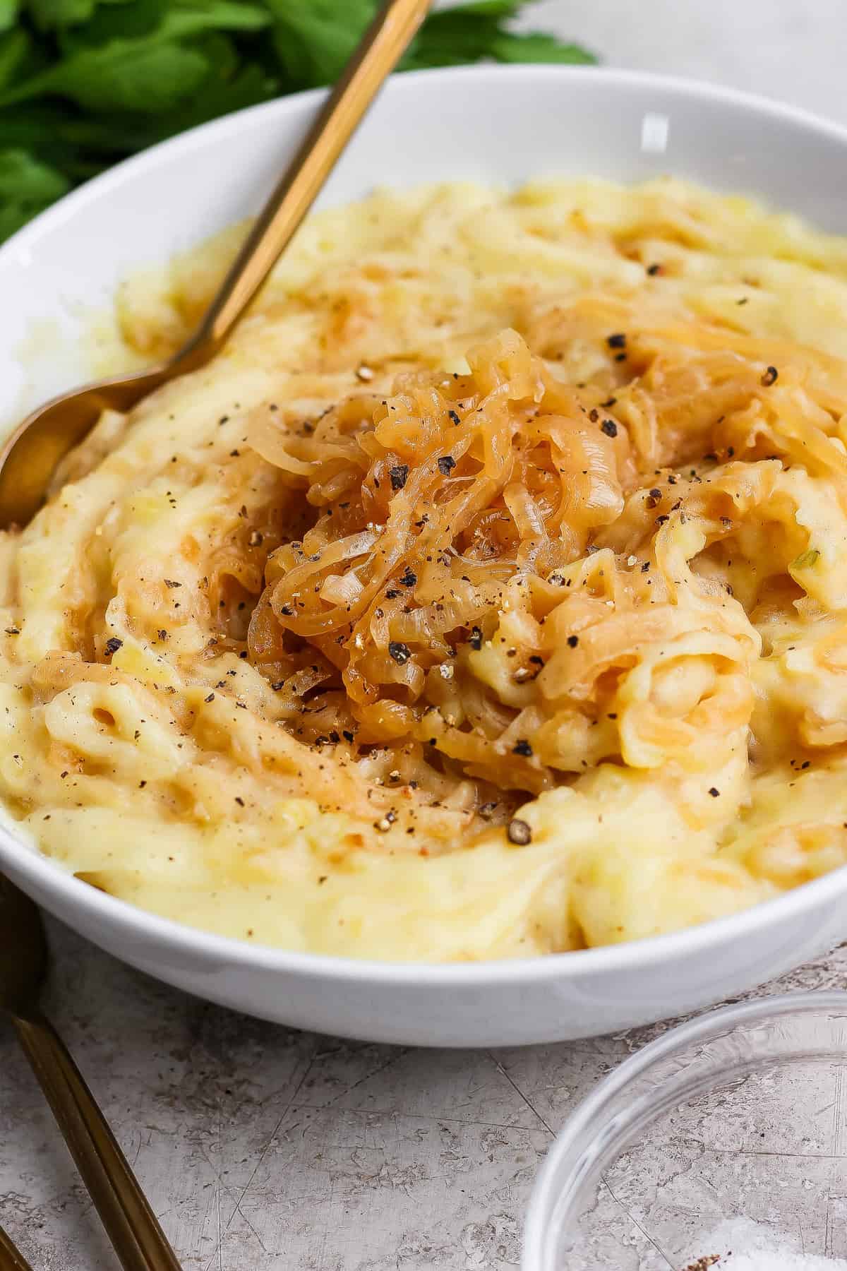 A bowl of mashed potatoes topped with caramelized onions and cracked black pepper, with a spoon resting inside.