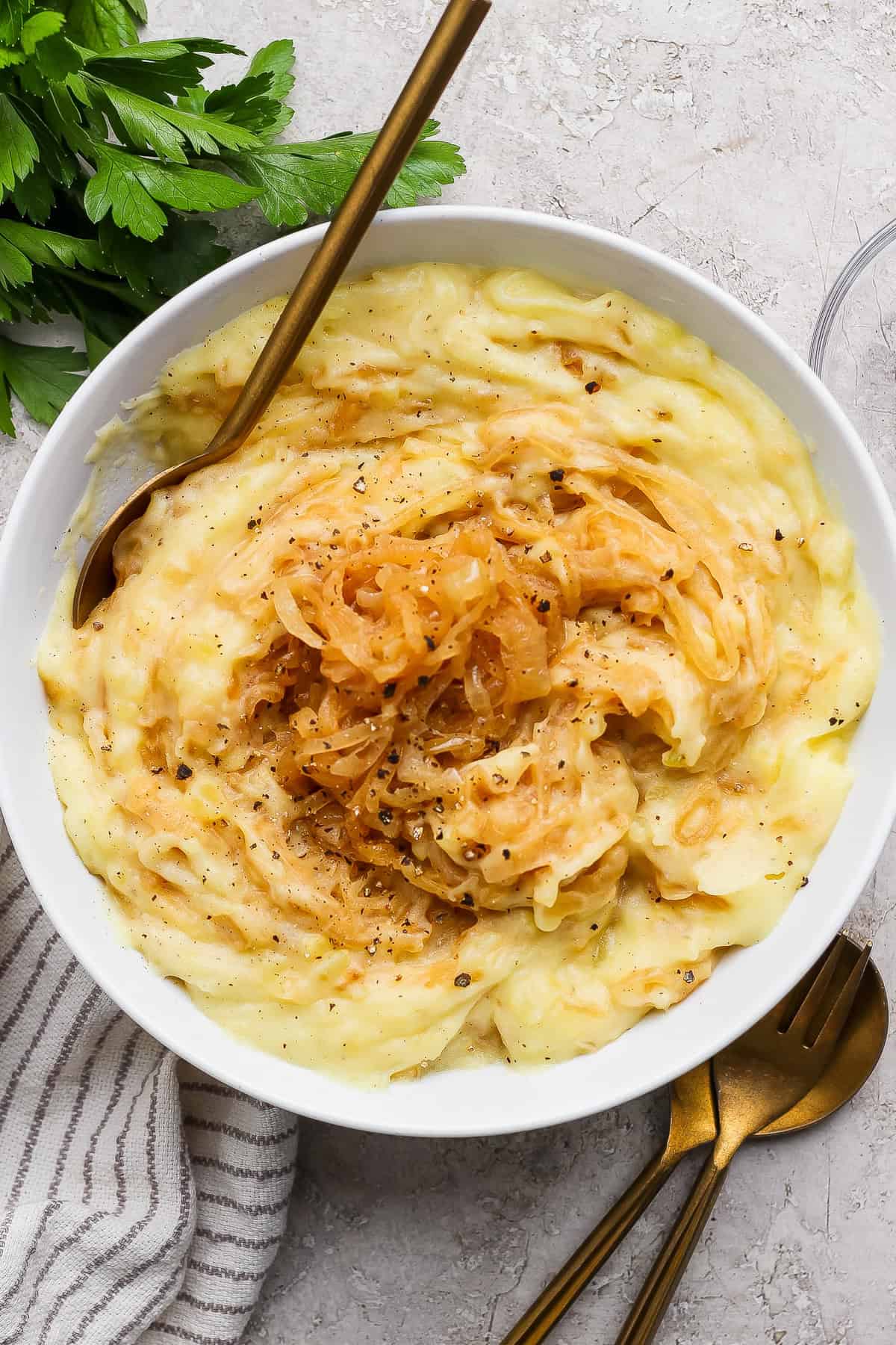 A bowl of mashed potatoes topped with caramelized onions and black pepper, with a gold fork and spoon beside the bowl on a light textured surface.