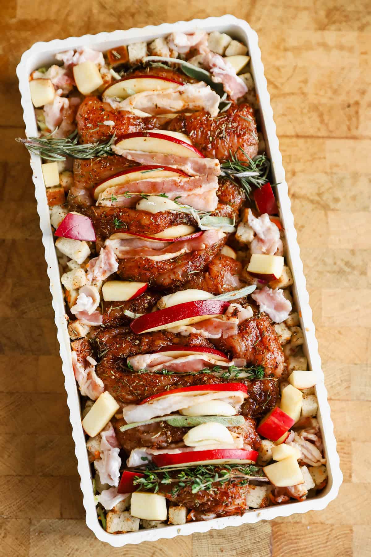 Raw seasoned chicken pieces topped with apple slices and herbs, arranged in a rectangular baking dish with cubed bread and chopped apples.