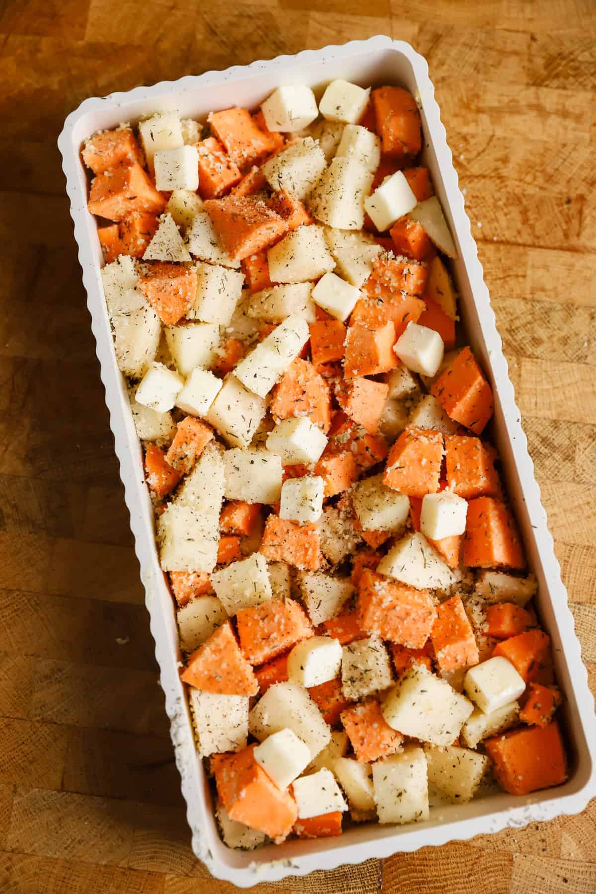 A rectangular baking dish filled with cubed sweet potatoes and white potatoes, topped with small cubes of butter and sprinkled with seasoning, on a wooden surface.