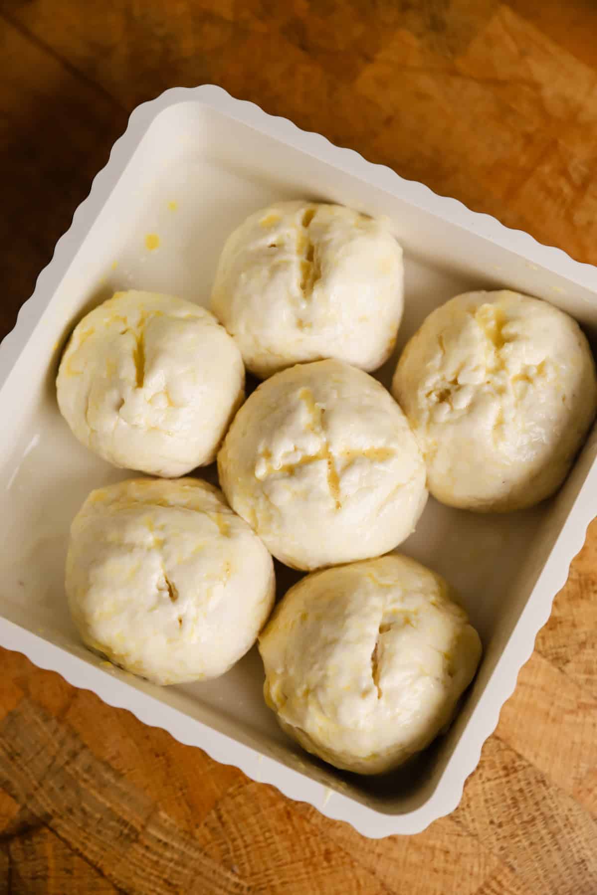 Six unbaked dough balls evenly arranged in a white baking dish, placed on a wooden surface.