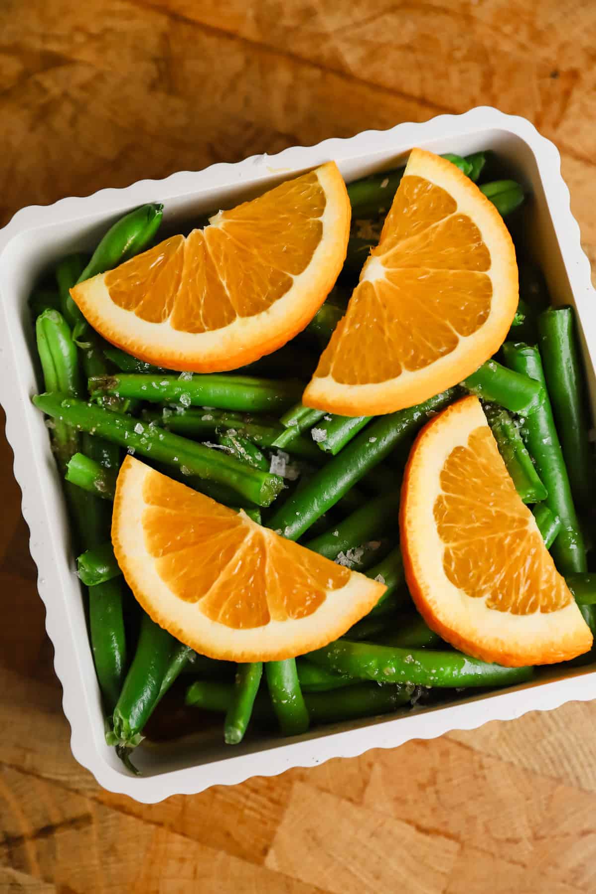 A white dish filled with green beans, topped with four orange slices, set on a wooden surface.