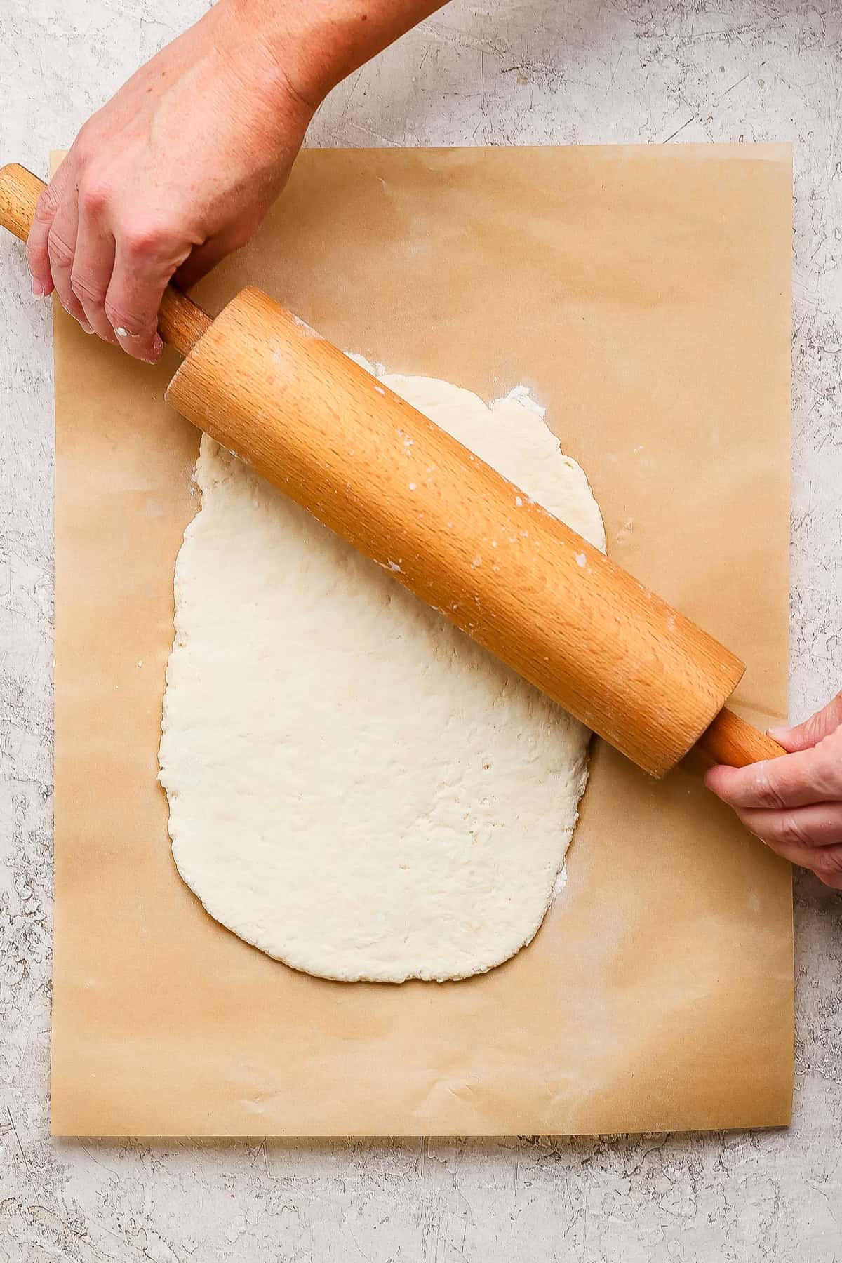 A person uses a wooden rolling pin to flatten dough on a sheet of parchment paper placed on a textured surface.