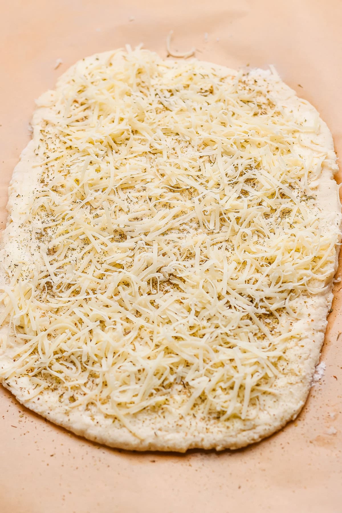 Unbaked flatbread dough topped with shredded cheese and herbs on a sheet of parchment paper.