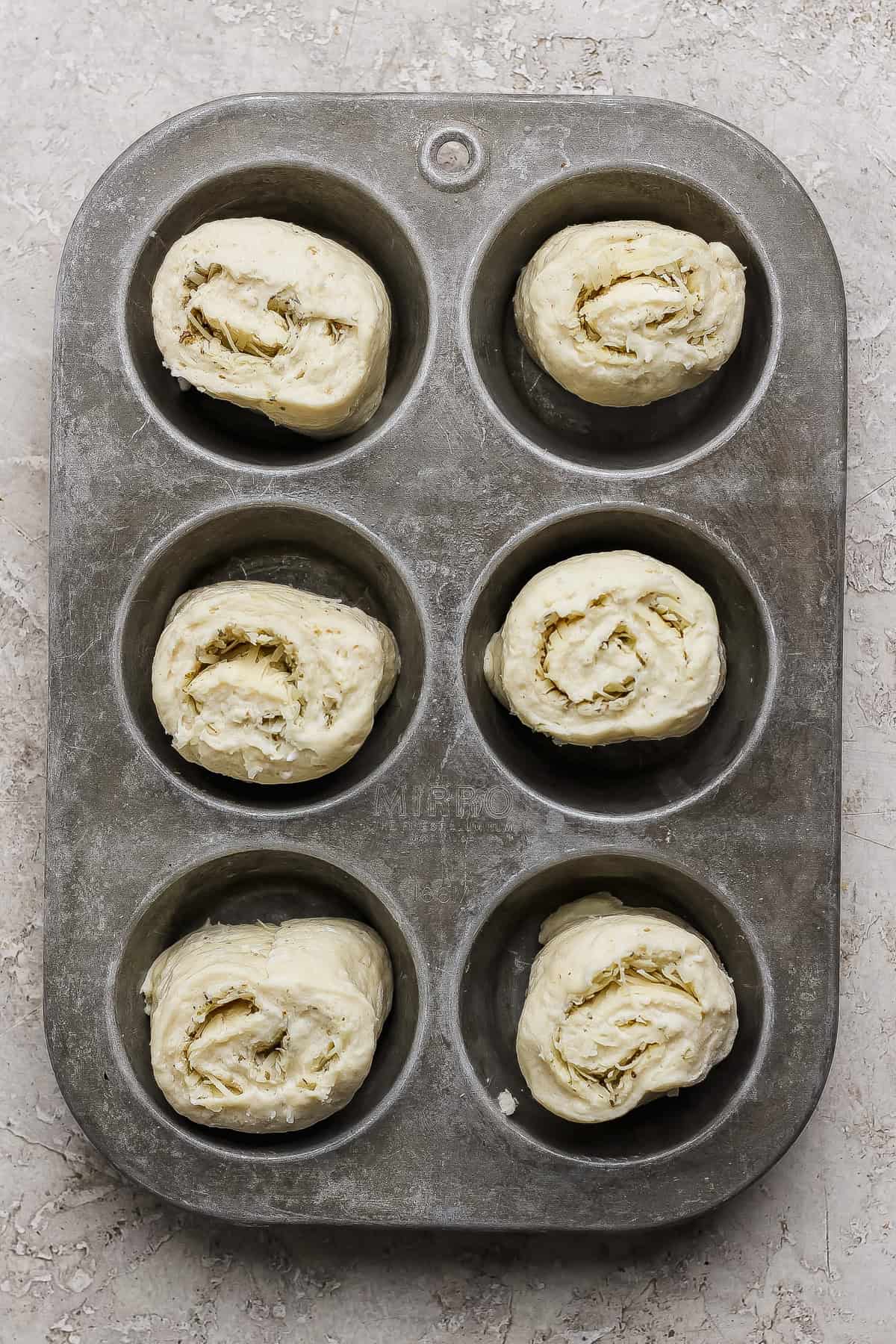 Six unbaked rolled dough pieces placed in a metal muffin tin, ready for baking, on a textured light surface.