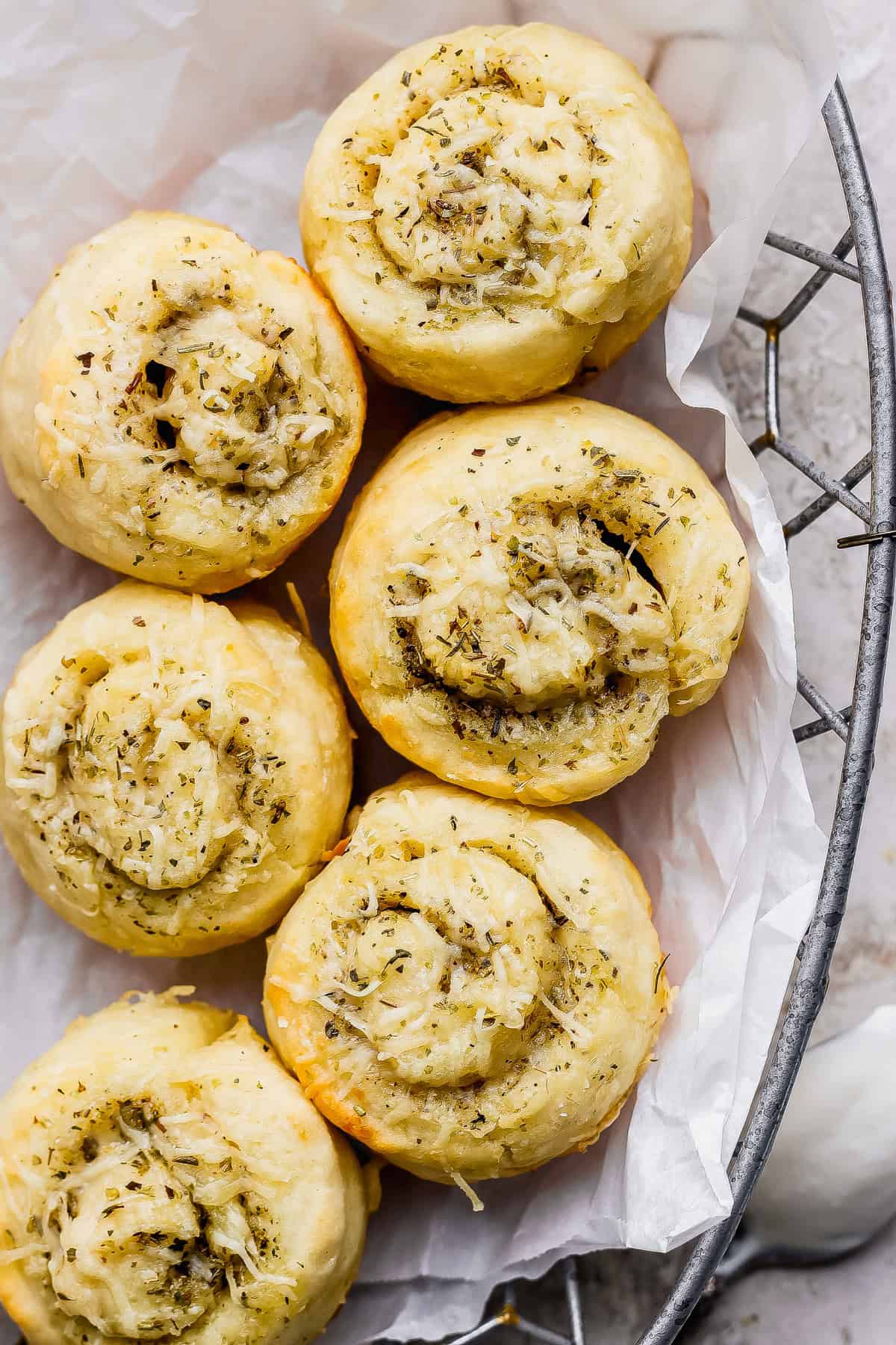 Six golden-brown, spiral-shaped bread rolls topped with herbs and shredded cheese, arranged on white parchment paper in a wire basket.