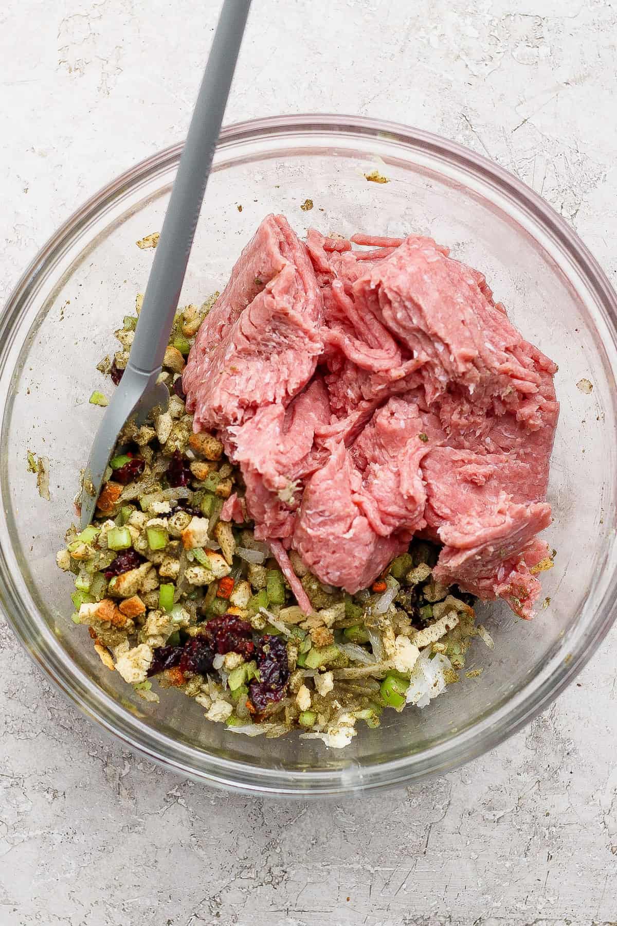 A glass bowl containing raw ground meat, diced vegetables, bread cubes, and a gray mixing spoon on a light countertop.