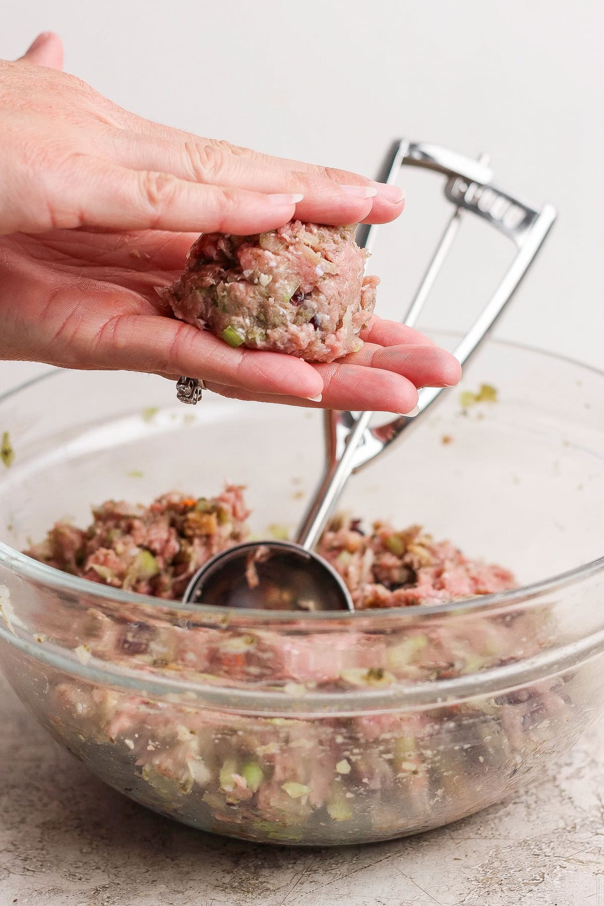 A hand shapes a portion of raw meat mixture into a ball over a glass bowl filled with more mixture; a metal scoop rests in the bowl.