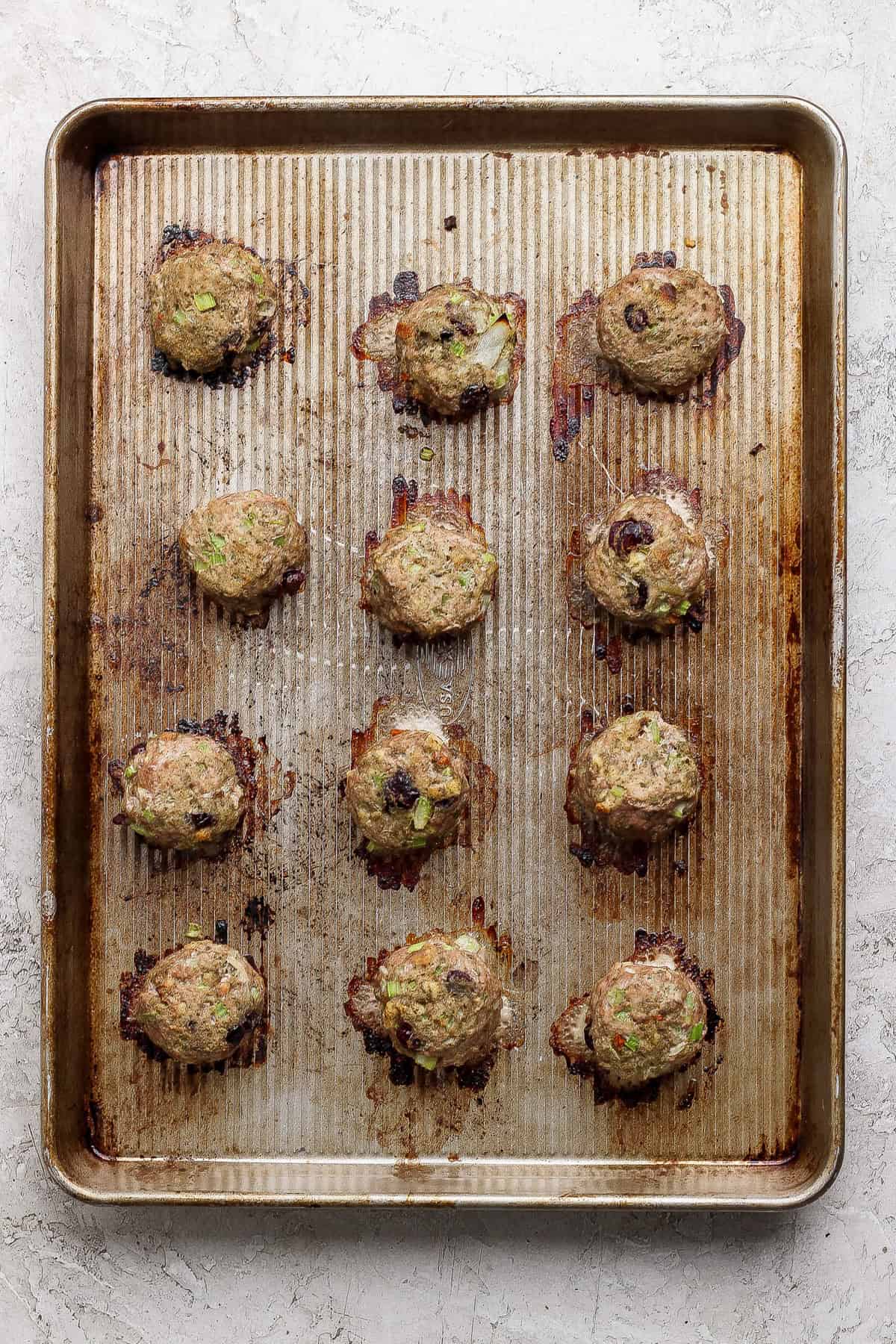 A baking sheet with twelve cooked meatballs, spaced apart, on a ridged metal tray with visible browning and juices.