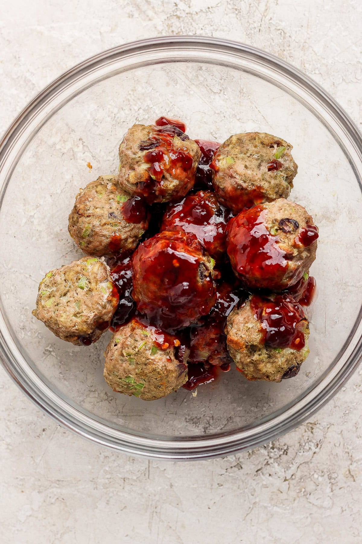 Meatballs coated with a dark sauce arranged in a glass bowl on a light textured surface.