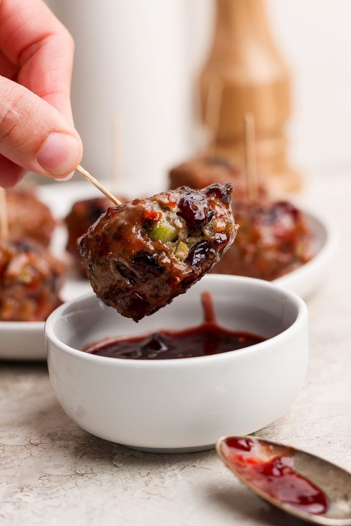 A hand holds a meatball on a toothpick, dipping it into a small bowl of dark sauce, with more meatballs and a spoon nearby.