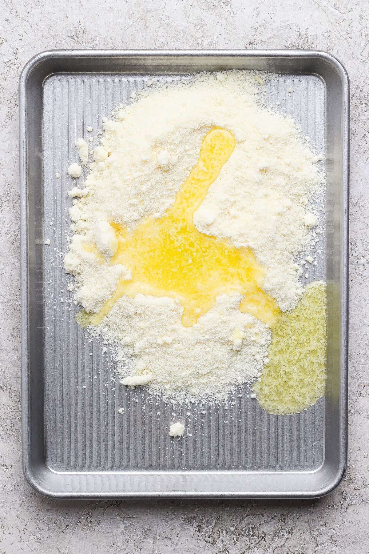 A baking sheet with grated cheese and melted butter spread across its surface.