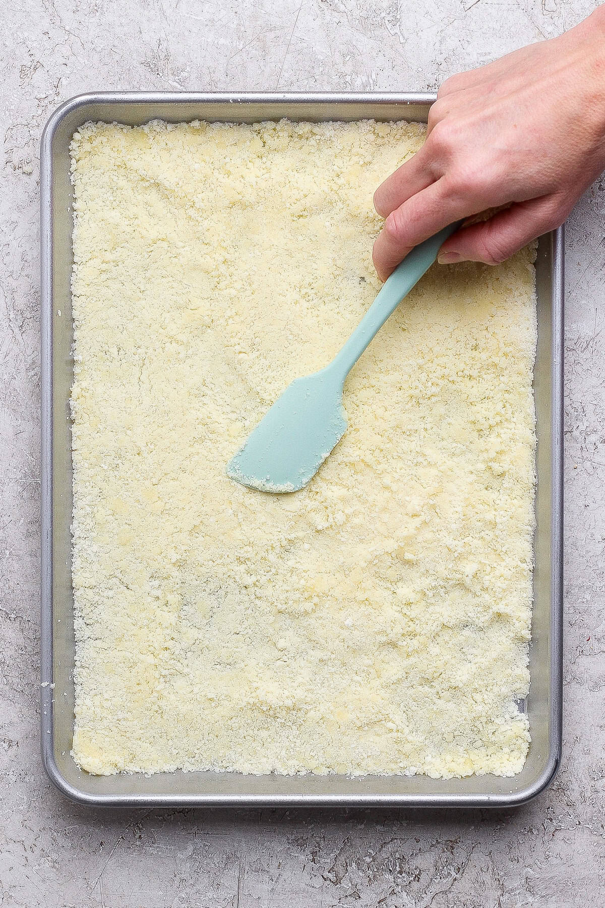 A hand uses a light blue spatula to smooth a layer of yellowish powder or crumbs on a rectangular baking sheet.