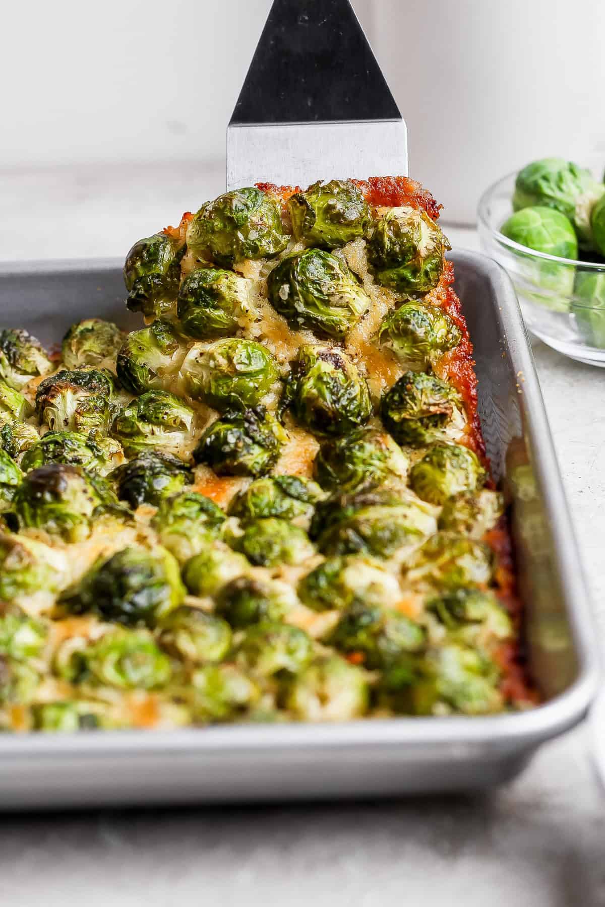 These roasted Brussel sprouts with parmesan are a great side dish staple for any time of year! All you need are 6 ingredients in order to make these crunchy, cheesy bites of heaven. A metal spatula lifts a piece of baked casserole topped with roasted brussels sprouts from a rectangular baking pan. A bowl of raw brussels sprouts sits in the background.