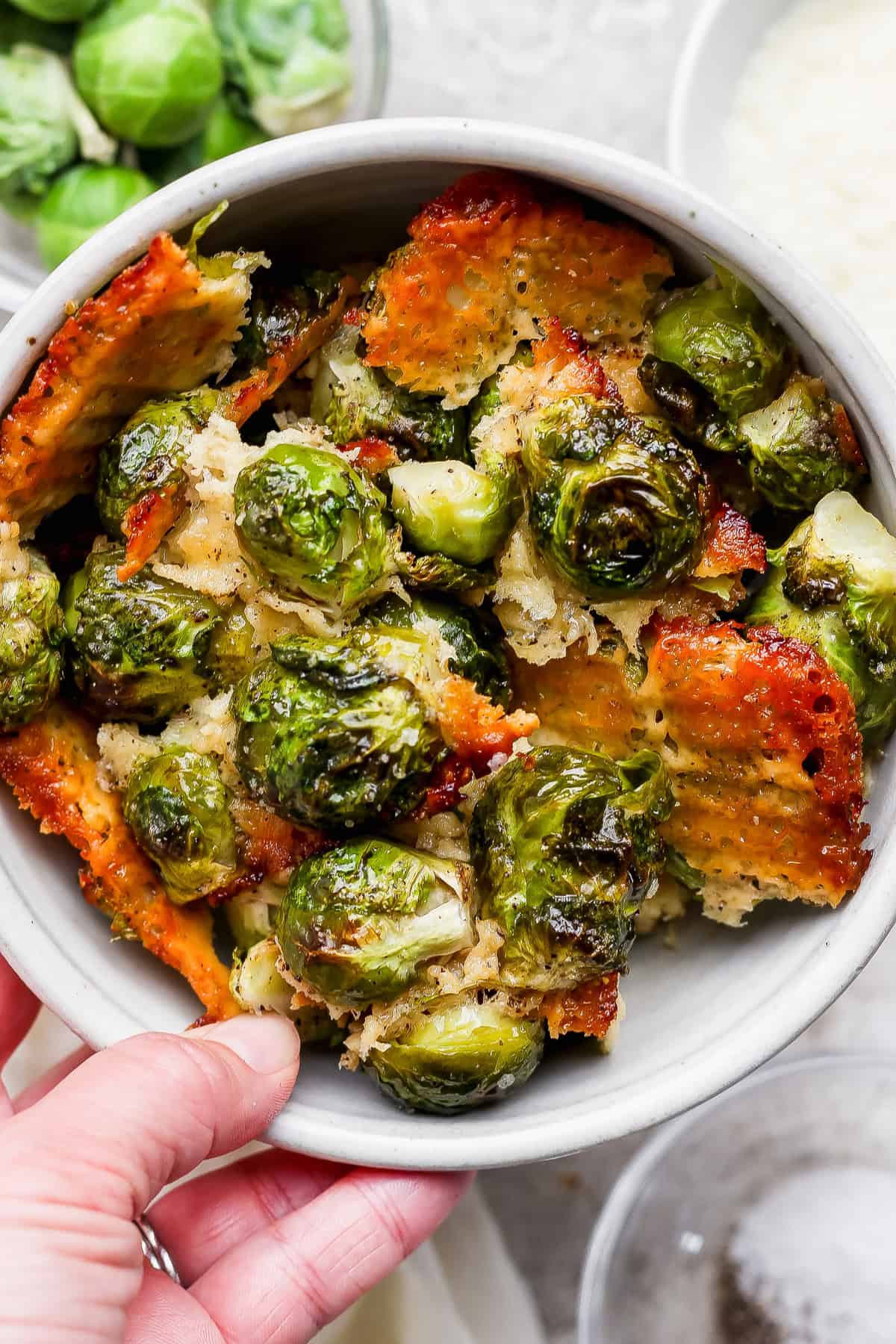 These roasted Brussel sprouts with parmesan are a great side dish staple for any time of year! All you need are 6 ingredients in order to make these crunchy, cheesy bites of heaven. A bowl of roasted Brussels sprouts topped with crispy, golden-brown cheese, with a hand holding the bowl.