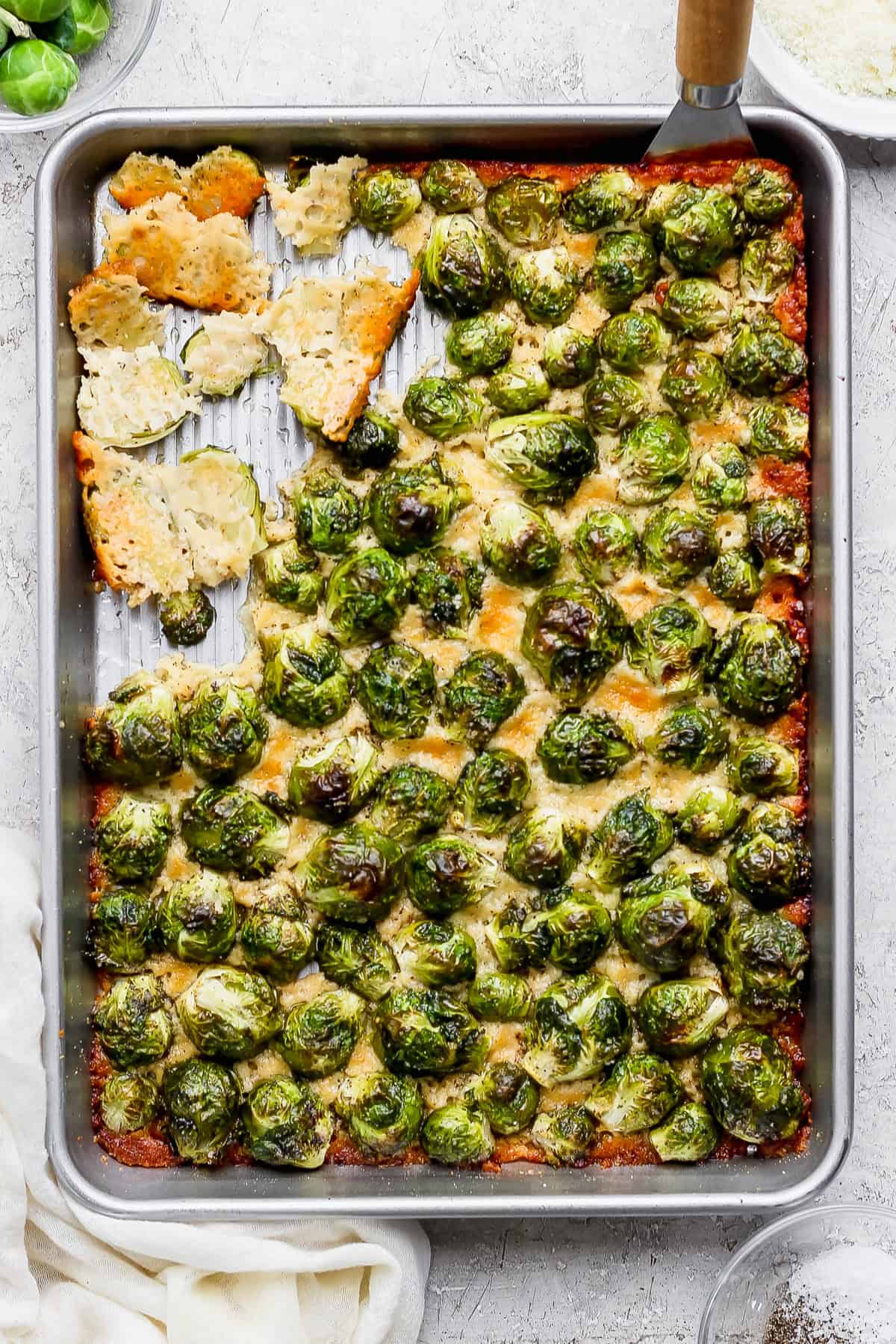 These roasted Brussel sprouts with parmesan are a great side dish staple for any time of year! All you need are 6 ingredients in order to make these crunchy, cheesy bites of heaven. A baking sheet with roasted Brussels sprouts topped with melted cheese, partially cut into squares with a spatula, on a light-colored surface.