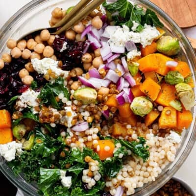 A vibrant couscous salad recipe featuring chickpeas, kale, roasted butternut squash, Brussels sprouts, Israeli couscous, red onion, dried cranberries, lentils, and crumbled cheese served with utensils.