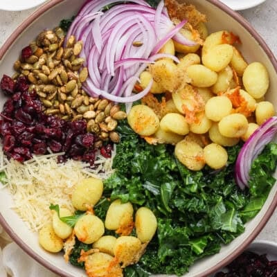 A bowl containing cooked gnocchi, kale, shredded parmesan, dried cranberries, pumpkin seeds, sliced red onions, and breadcrumbs.